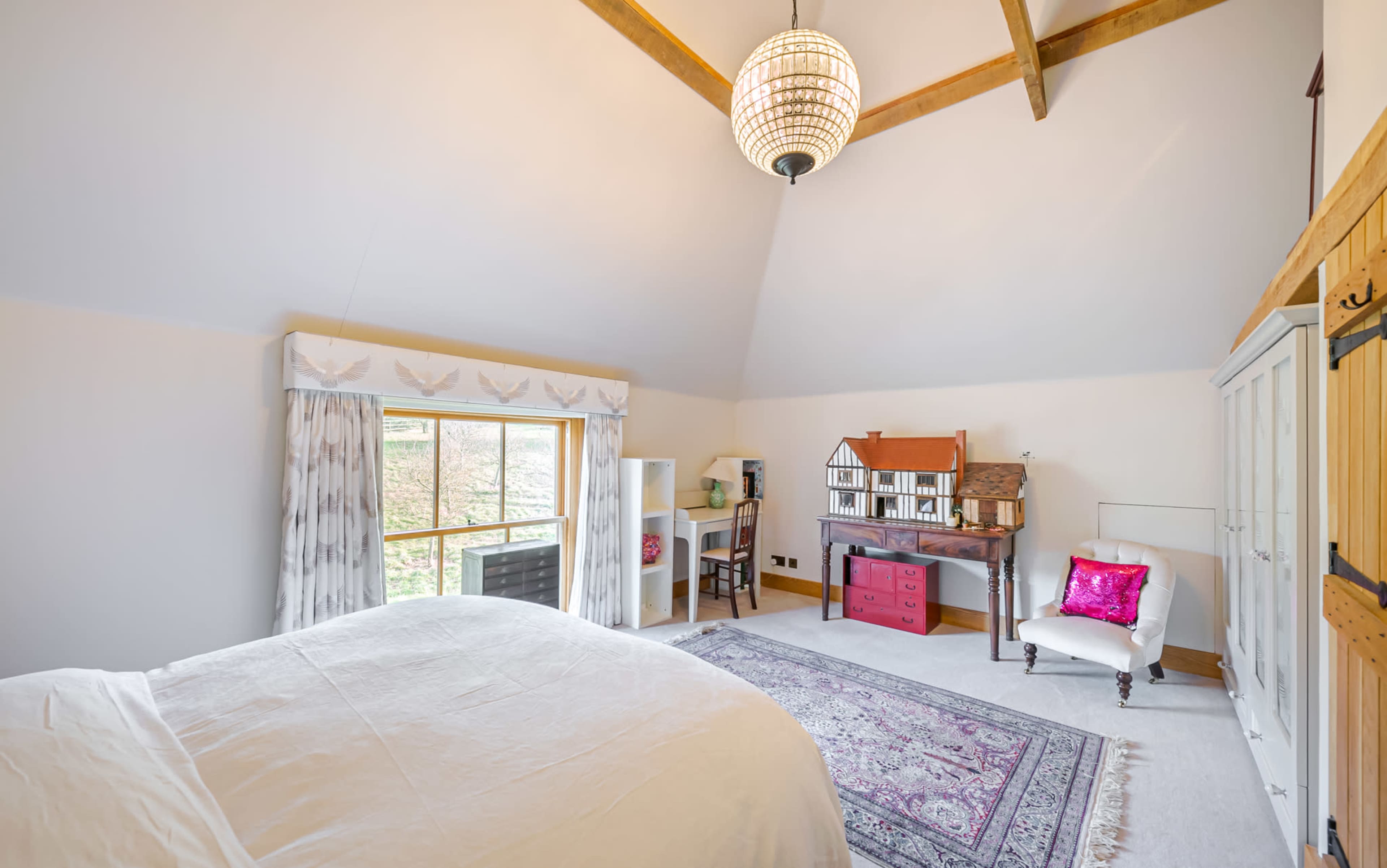 A spacious bedroom features a large window, a bed with white linens, a desk with a house model, and a white armchair.