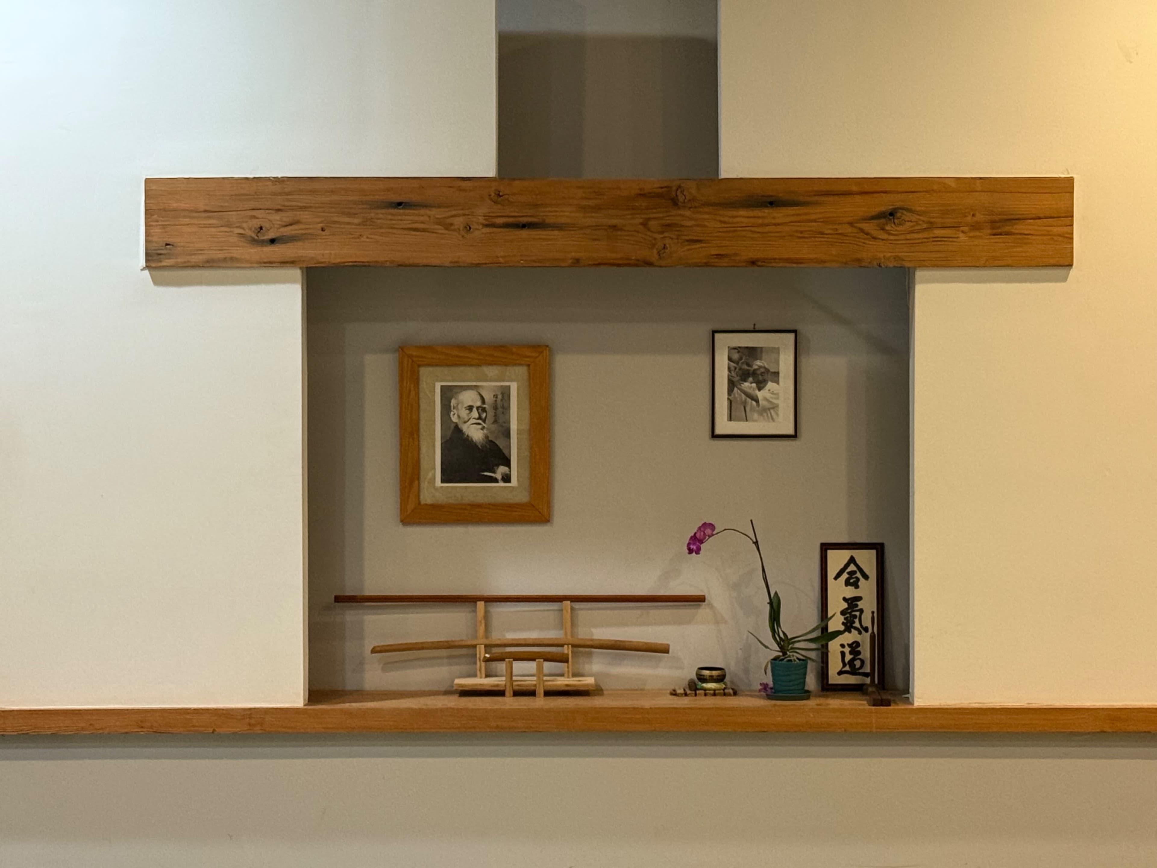 The image features a minimalist alcove with a wooden shelf, framed photographs, a decorative bamboo sword display, and a potted orchid.