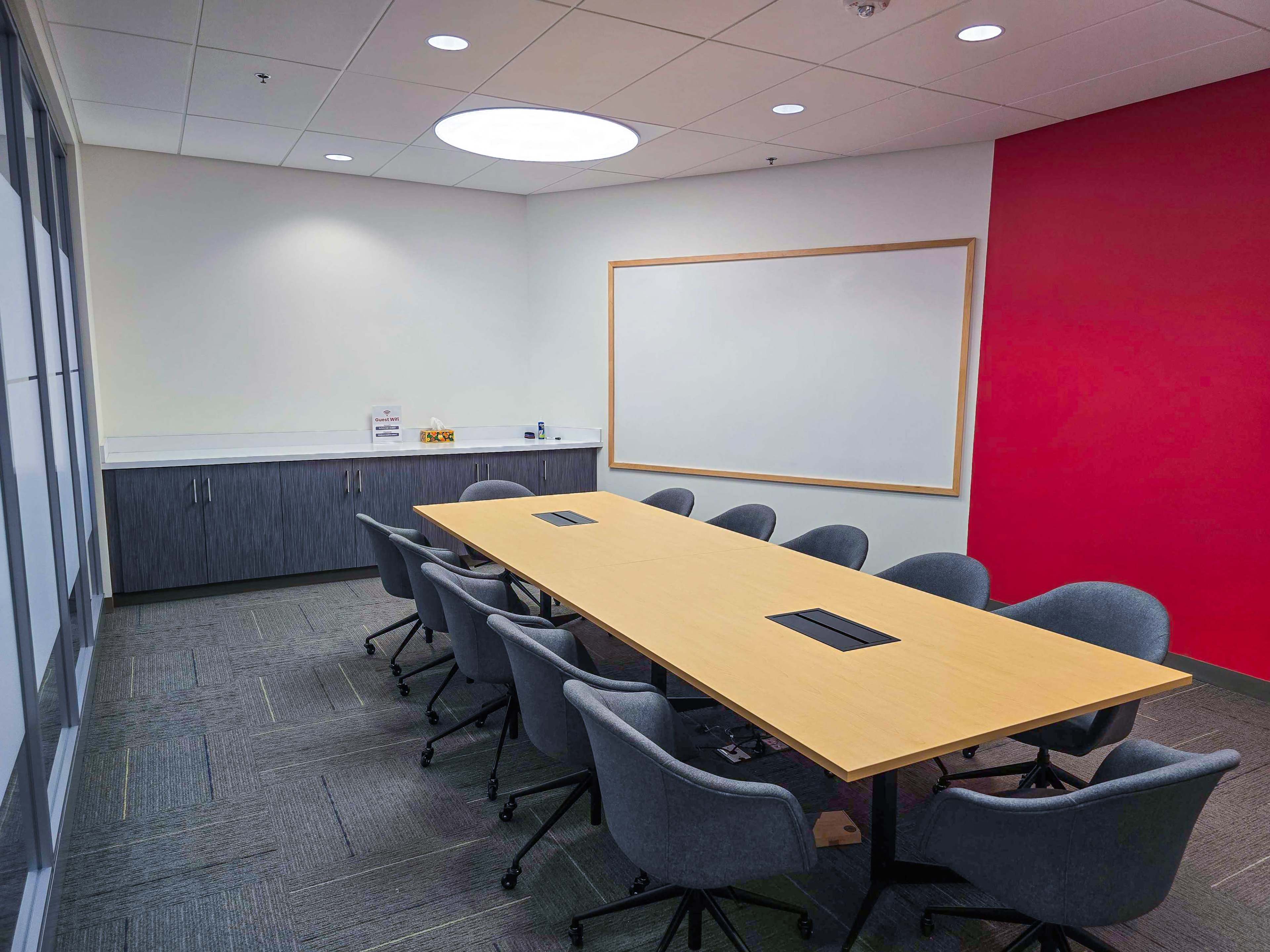 The image shows a conference room with a large rectangular table surrounded by ergonomic chairs, a whiteboard on the wall, and cabinets on the side.