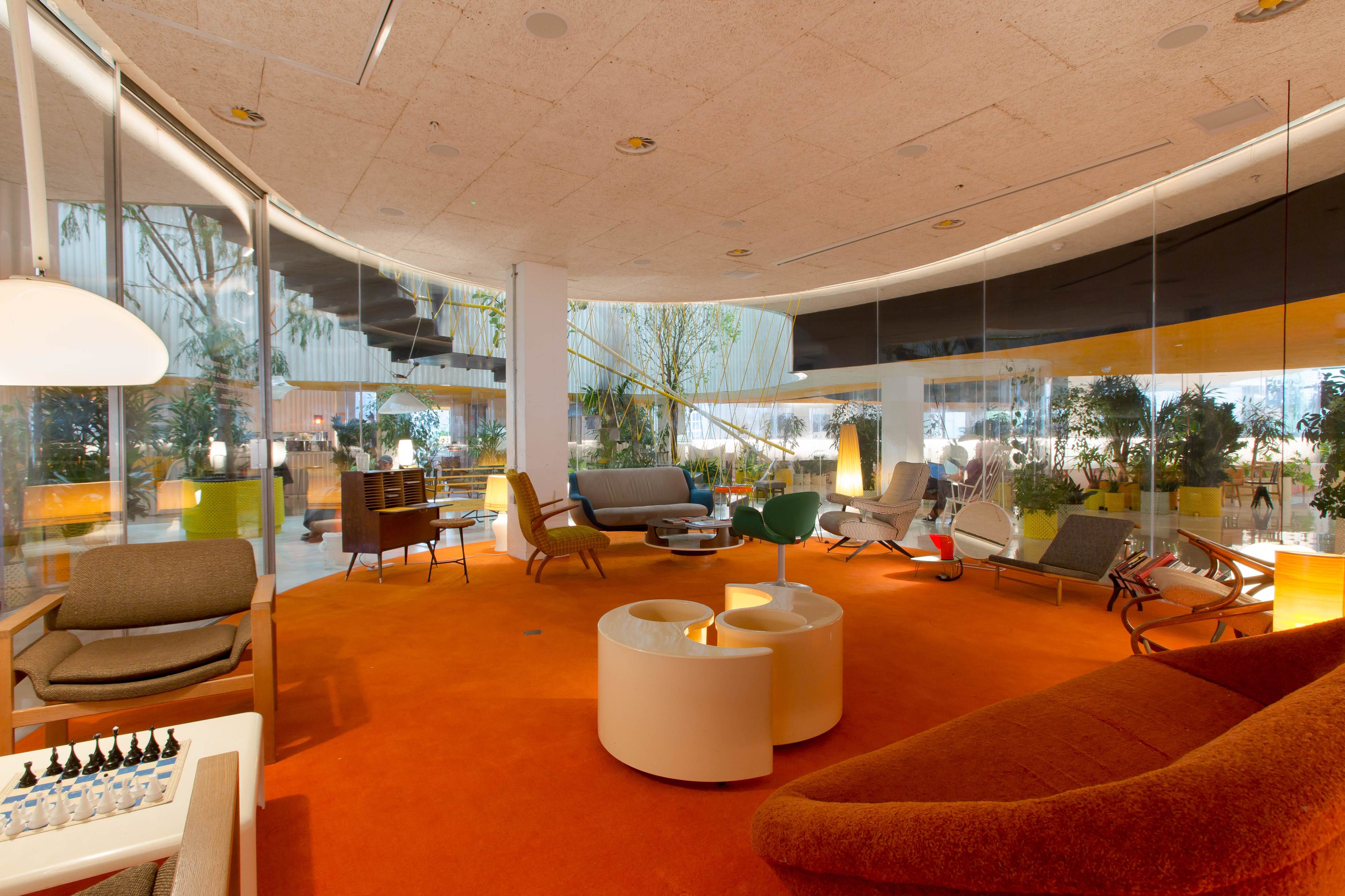 The image shows a modern lounge area featuring various seating arrangements, large windows with greenery outside, and an orange carpet.