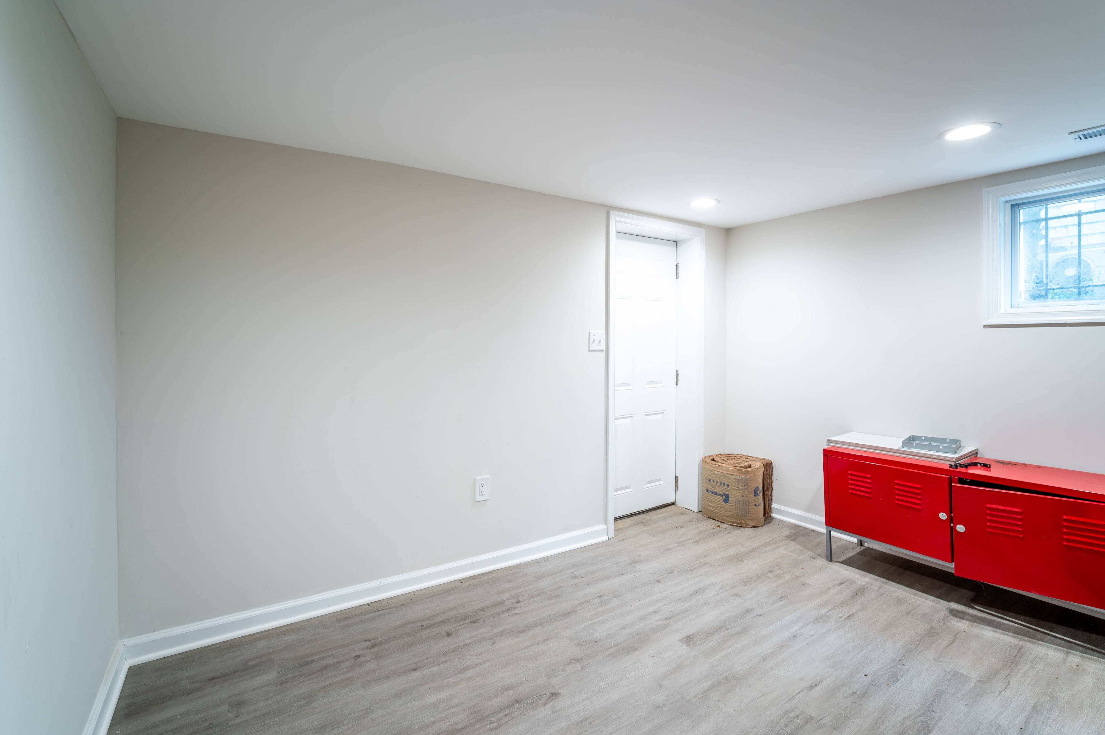 A sparsely furnished basement room with light-colored walls, a door on the right, and a red storage cabinet against one wall.