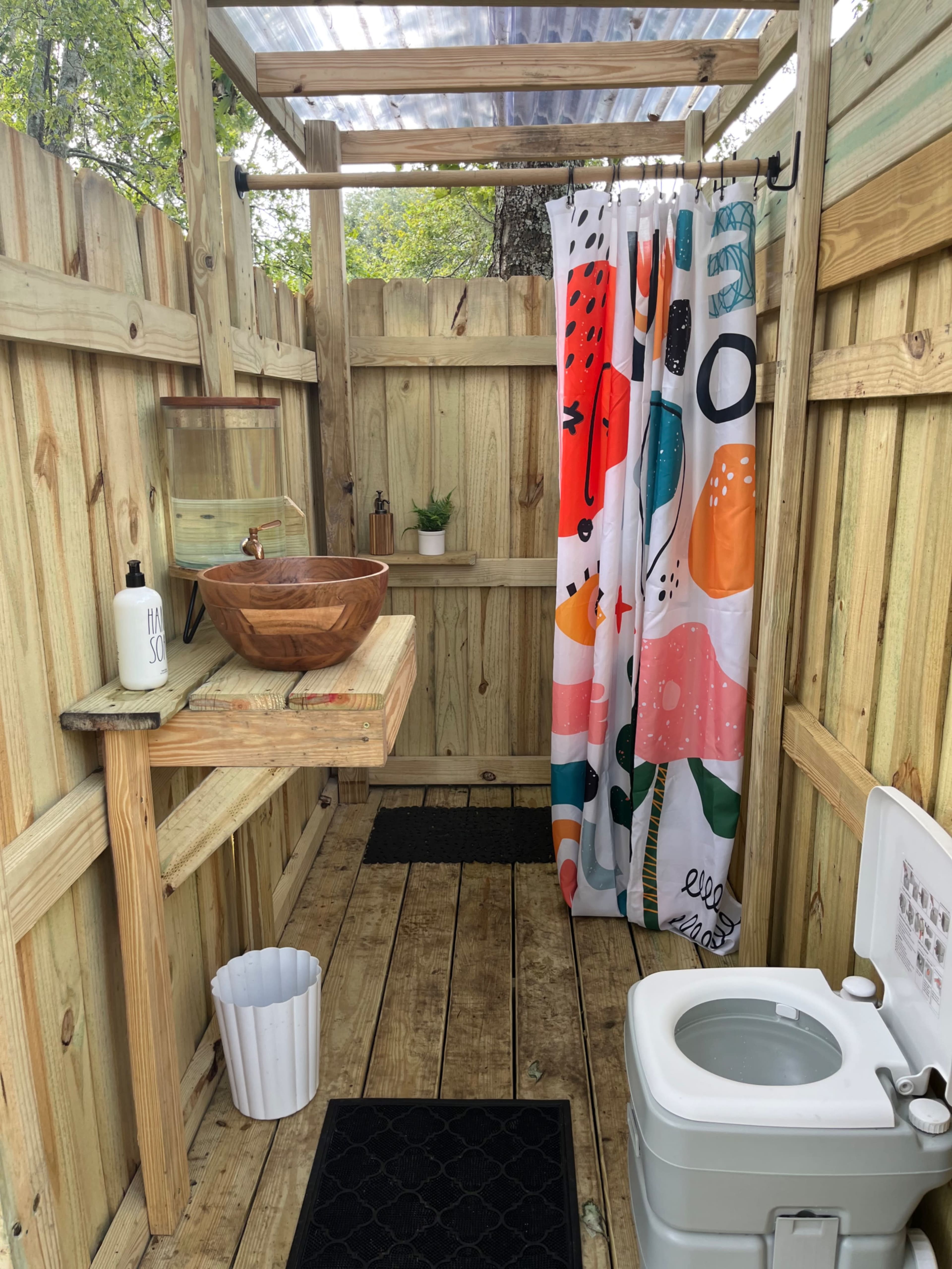 An outdoor shower area with wooden walls, a wooden sink with a bowl, a colorful shower curtain, and a portable toilet.