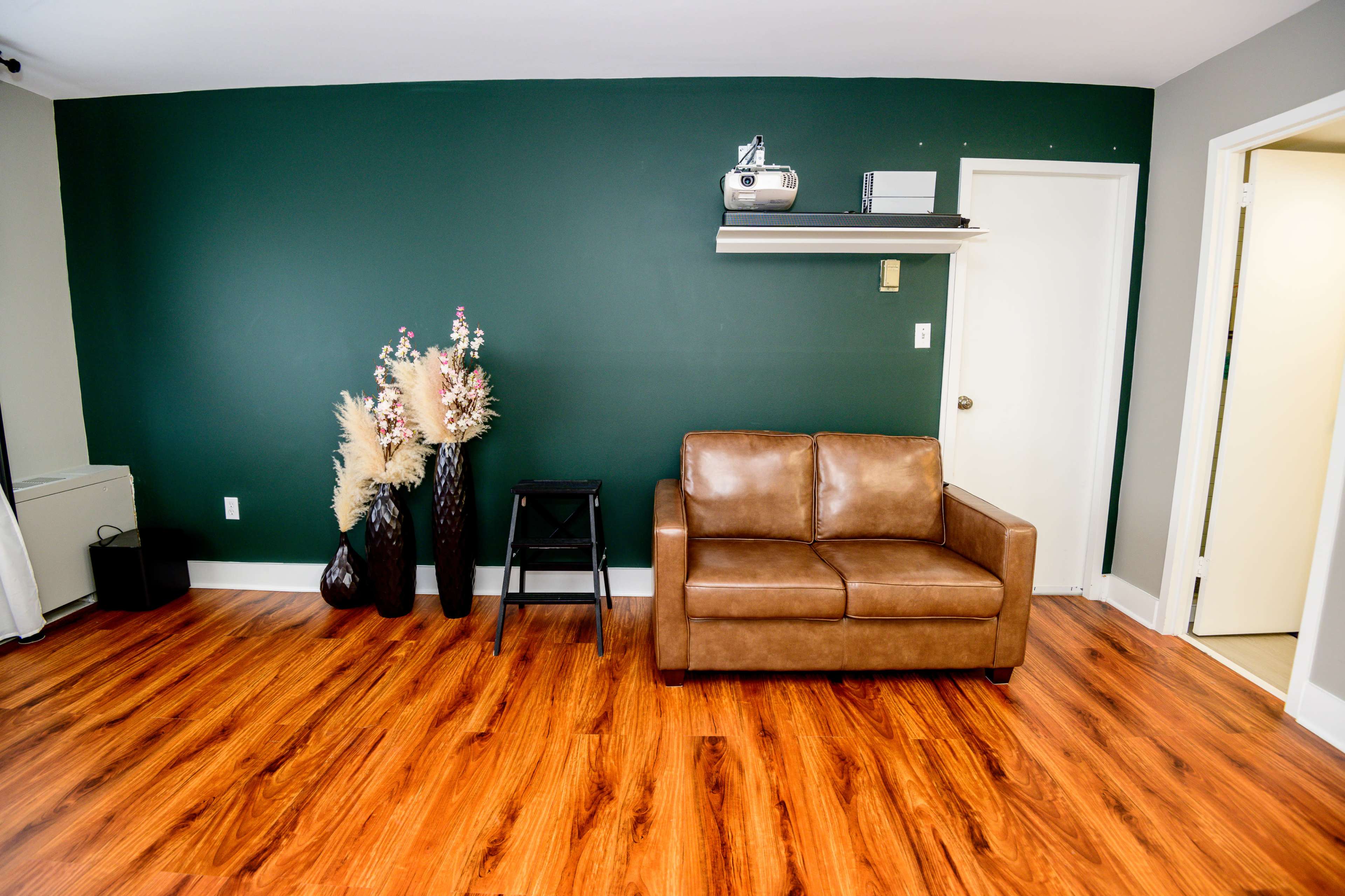 A small room with a brown leather couch, a black stool, decorative vases with dried flowers, and a green accent wall.