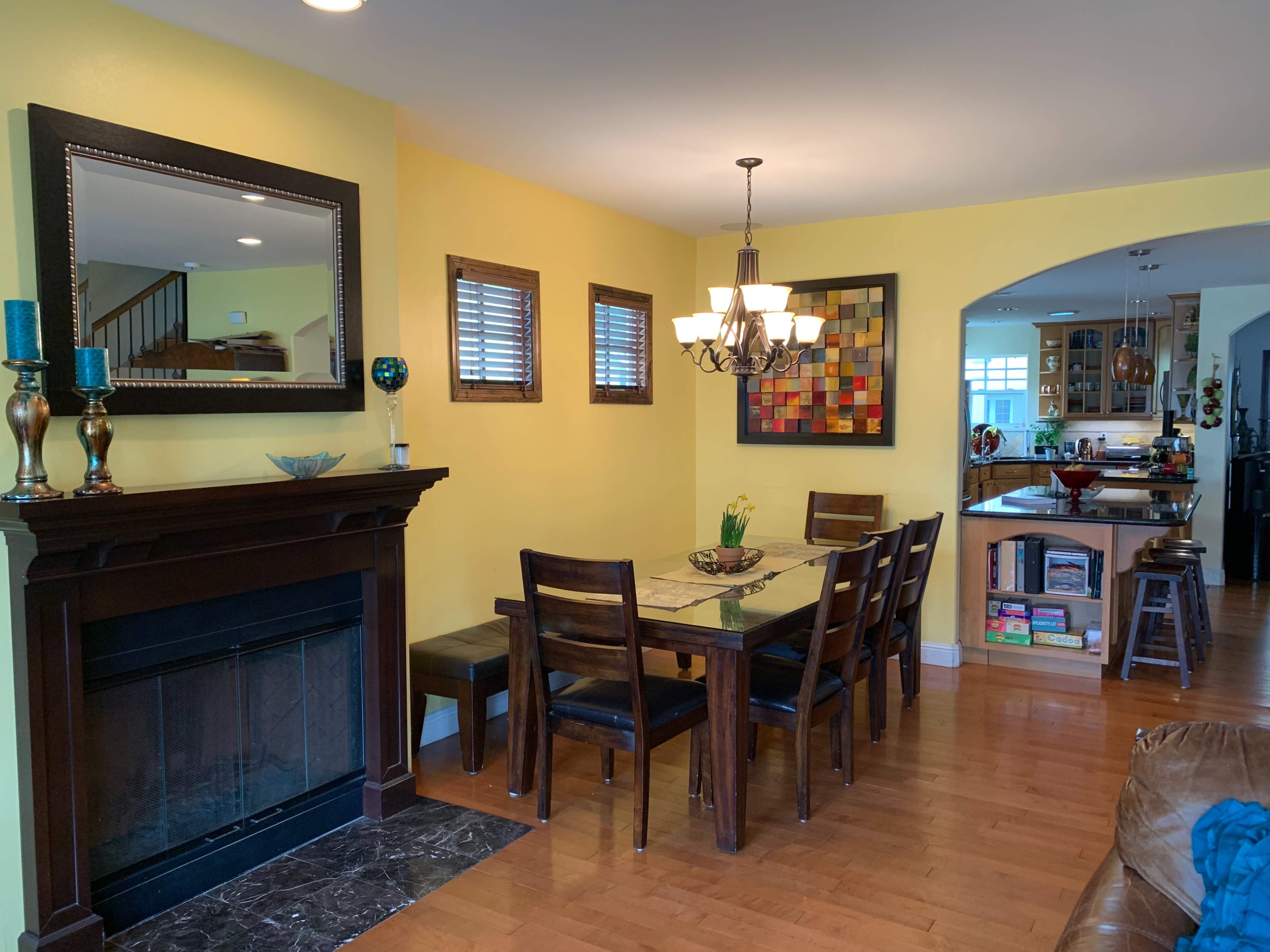 A dining area features a wooden table with six chairs, a fireplace, and yellow walls adorned with a mirror and a colorful artwork.