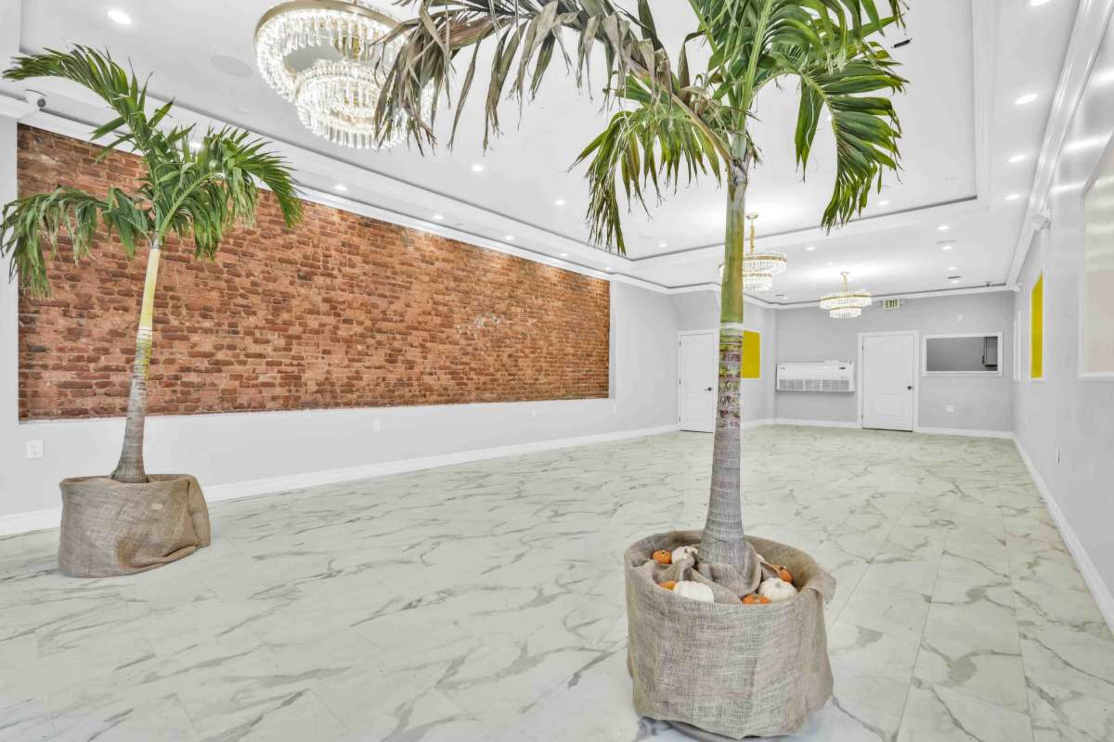 The room features a marble floor, two potted palm trees, and a textured brick wall with chandeliers above.