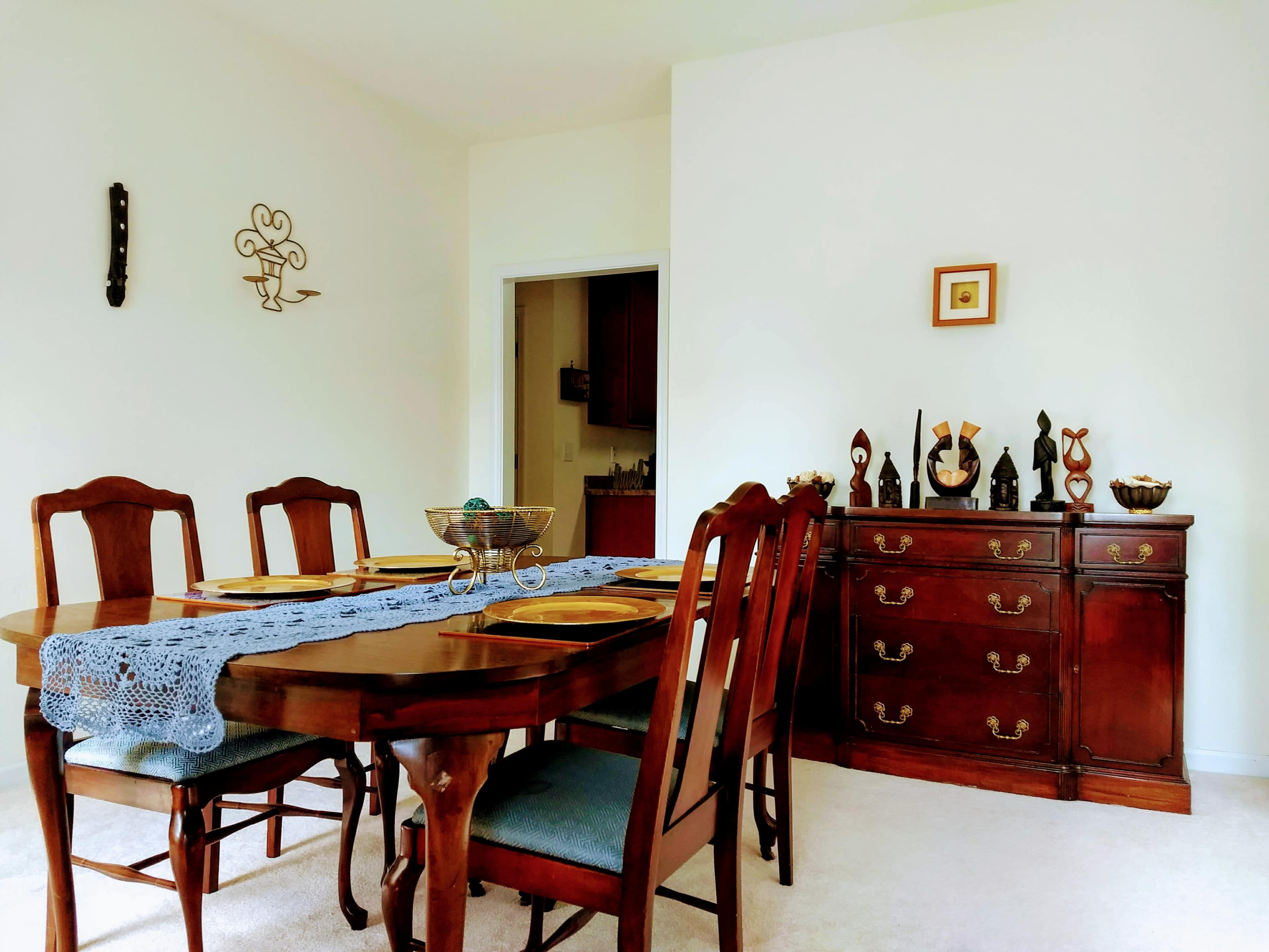 A wooden dining table is set with plates and a blue table runner, surrounded by chairs, adjacent to a sideboard decorated with various sculptures and tableware.
