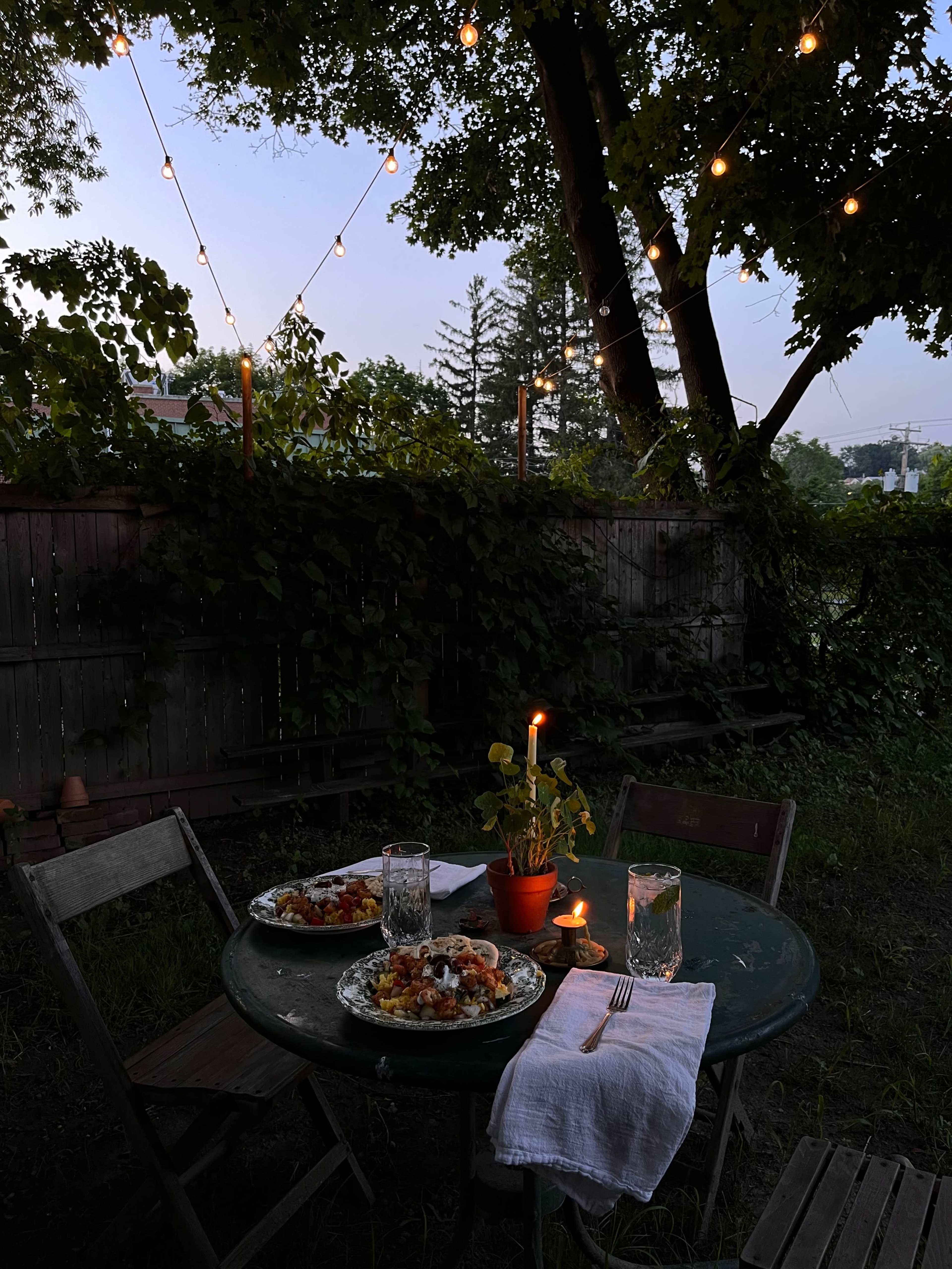 A small round table is set for two with plates of food, glasses of water, and a candle in a potted plant, beneath string lights illuminating a backyard at dusk.
