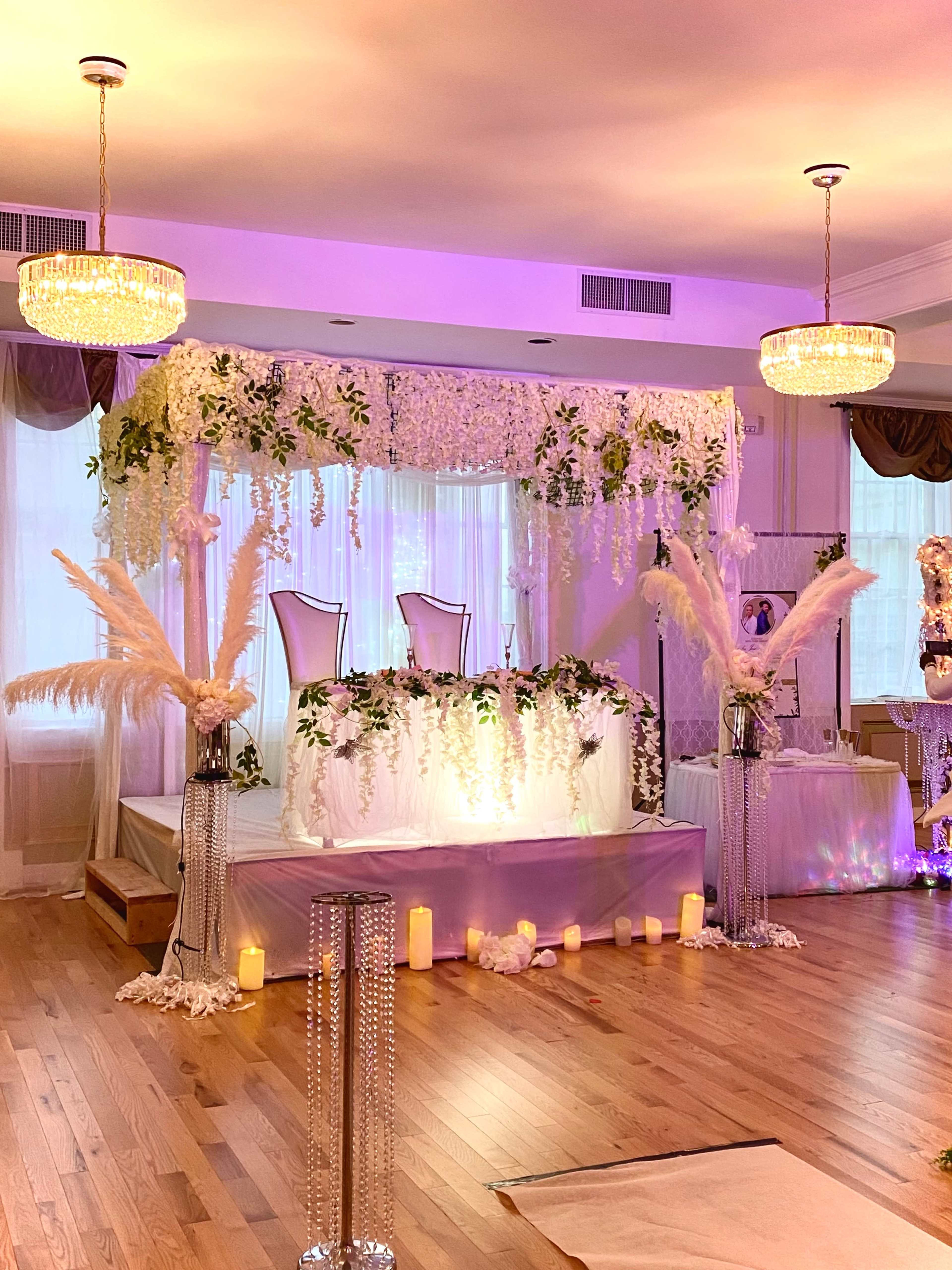 A decorated event space featuring a floral arch, a raised platform with two chairs, and soft lighting accentuated by pendant chandeliers.