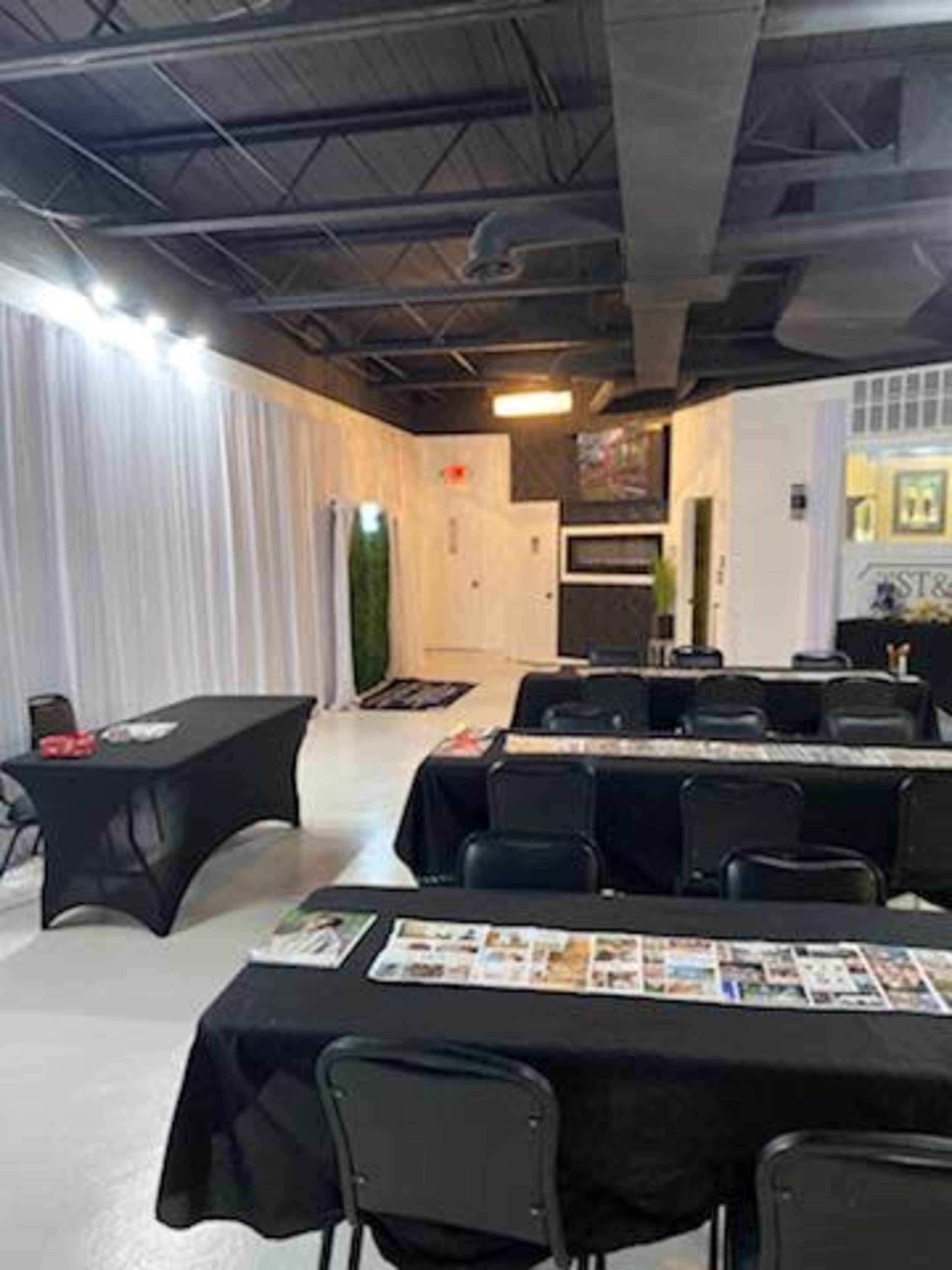The image shows an indoor event space with several rows of black tables and chairs, partially decorated with white drapes and a large wall-mounted screen.