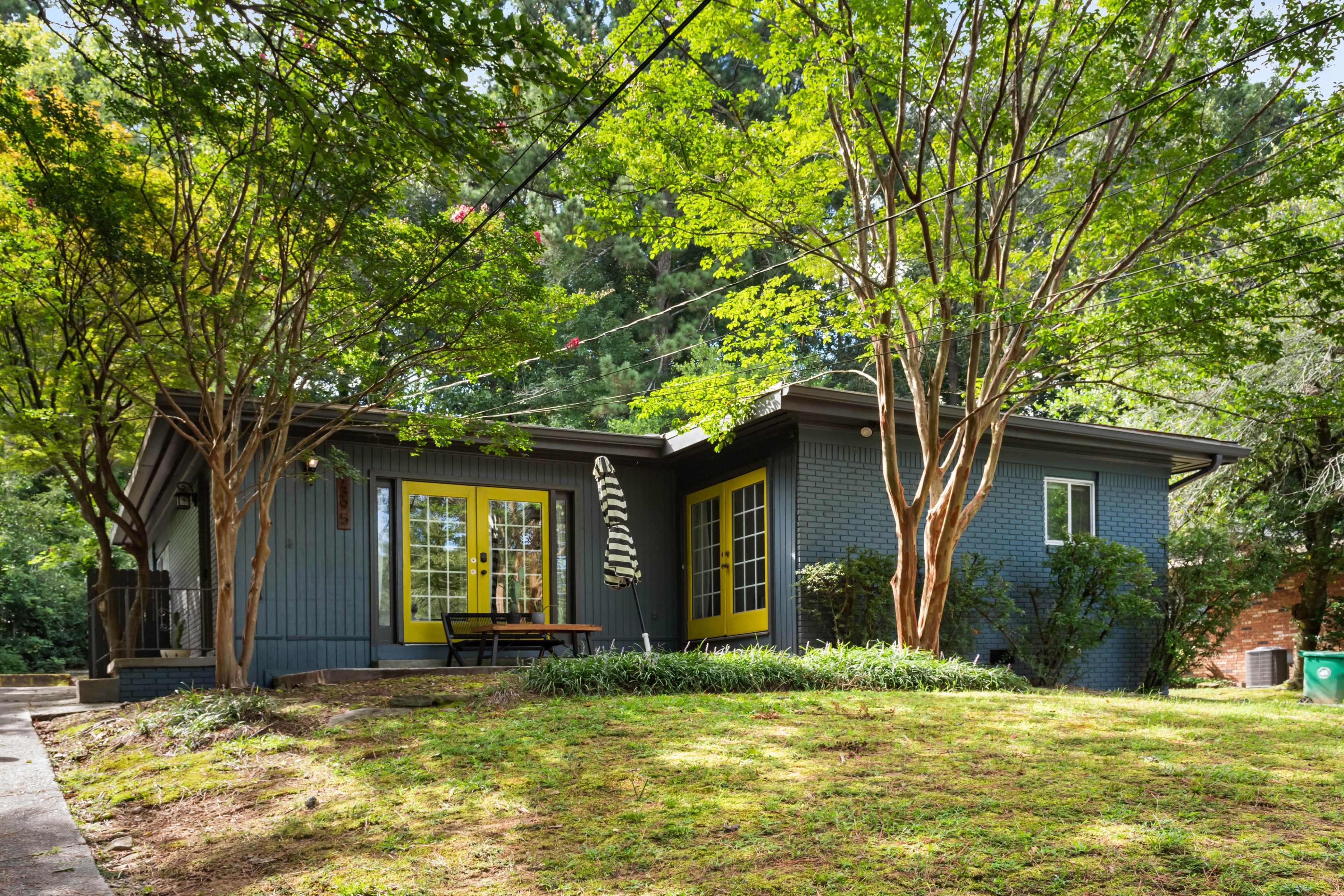 A modern gray house with bright yellow accents is surrounded by green trees and a manicured lawn.