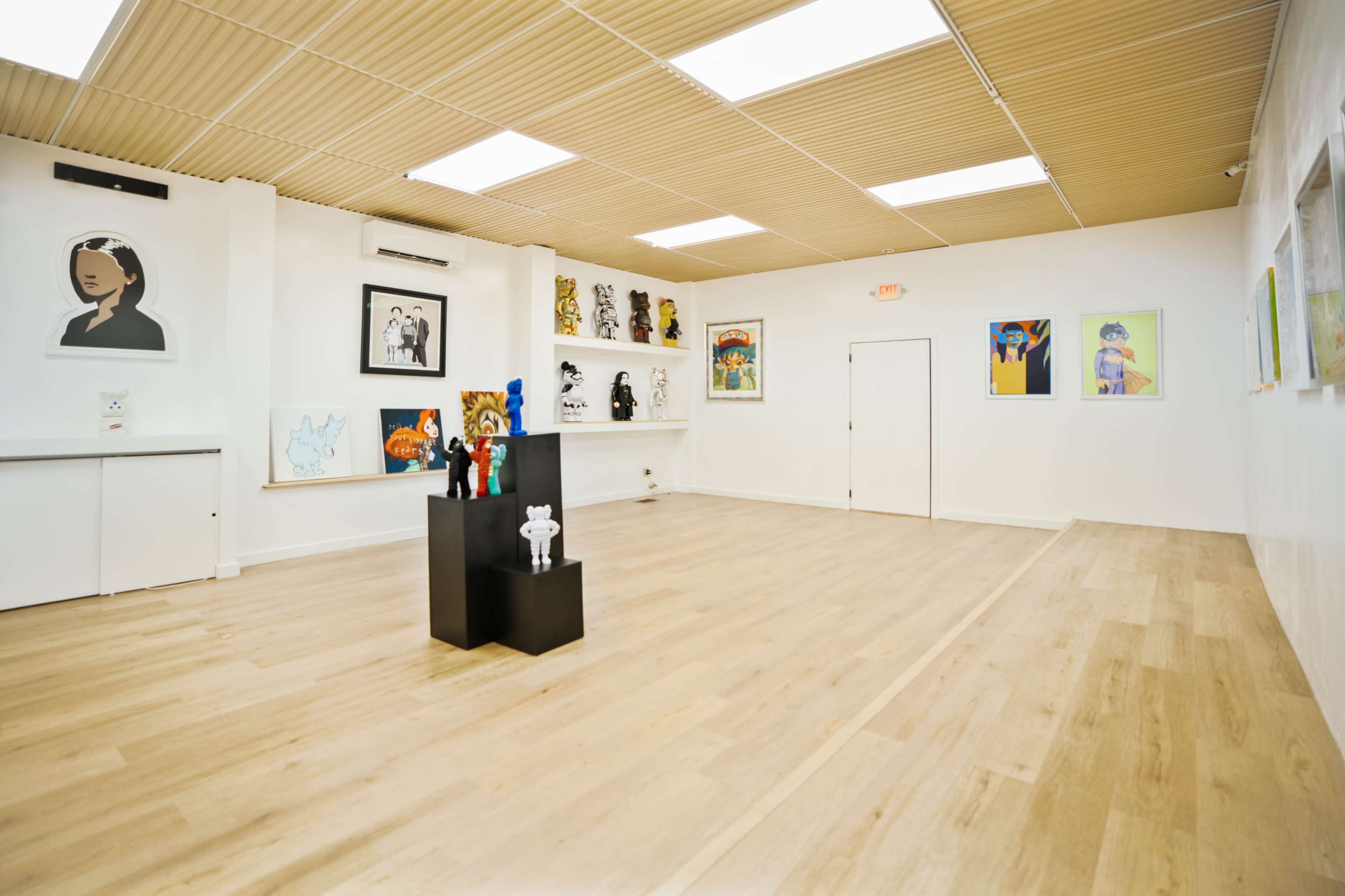 An empty art gallery with wooden flooring, white walls featuring various artworks, and a central display stand.