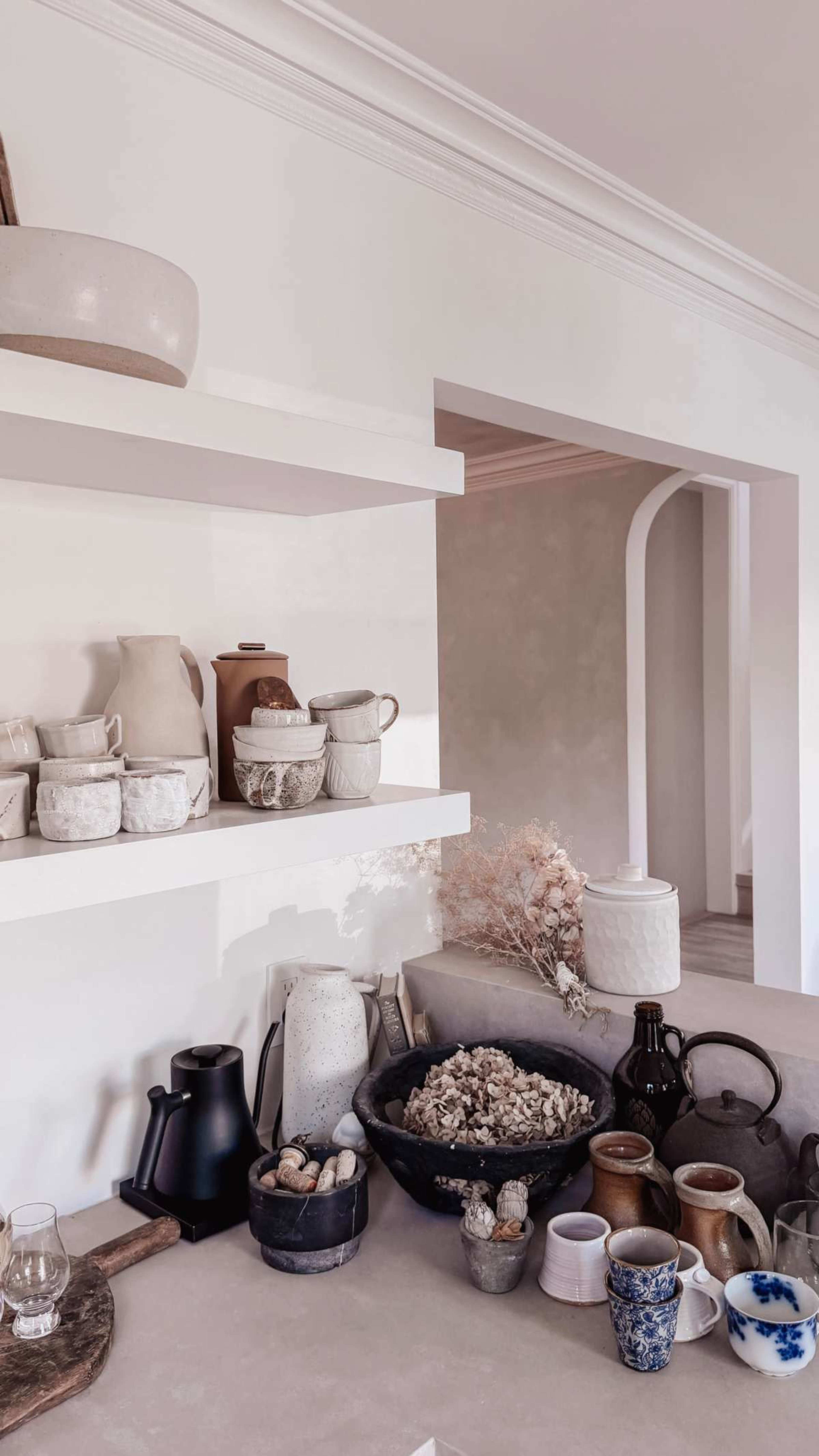 The image shows a minimalist kitchen shelf with various ceramic pots and jars arranged neatly, alongside a countertop decorated with a bowl of natural elements and additional mugs.