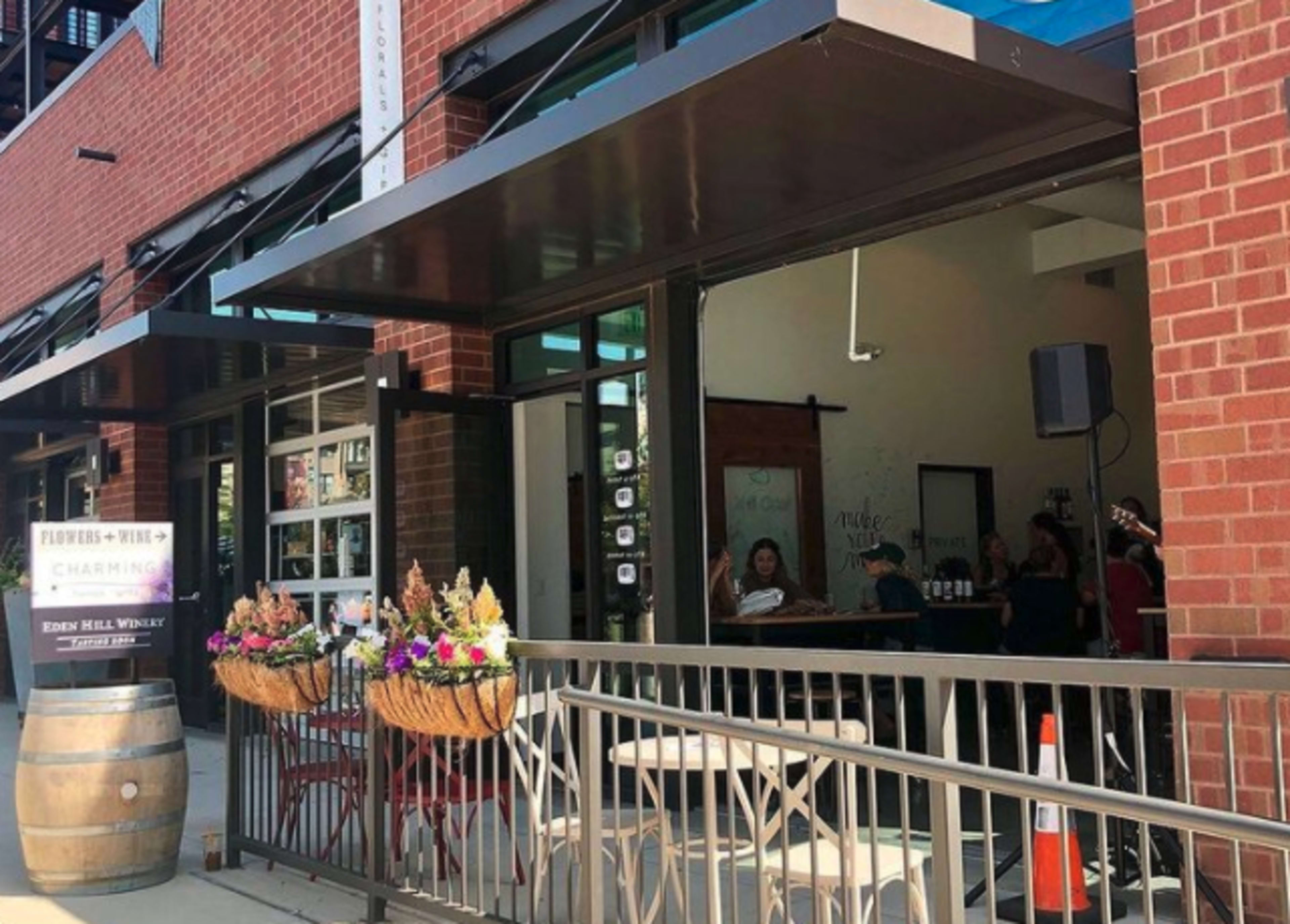 A storefront with a canopy features outdoor seating and flower baskets, alongside a sign for charging stations and a wooden barrel.