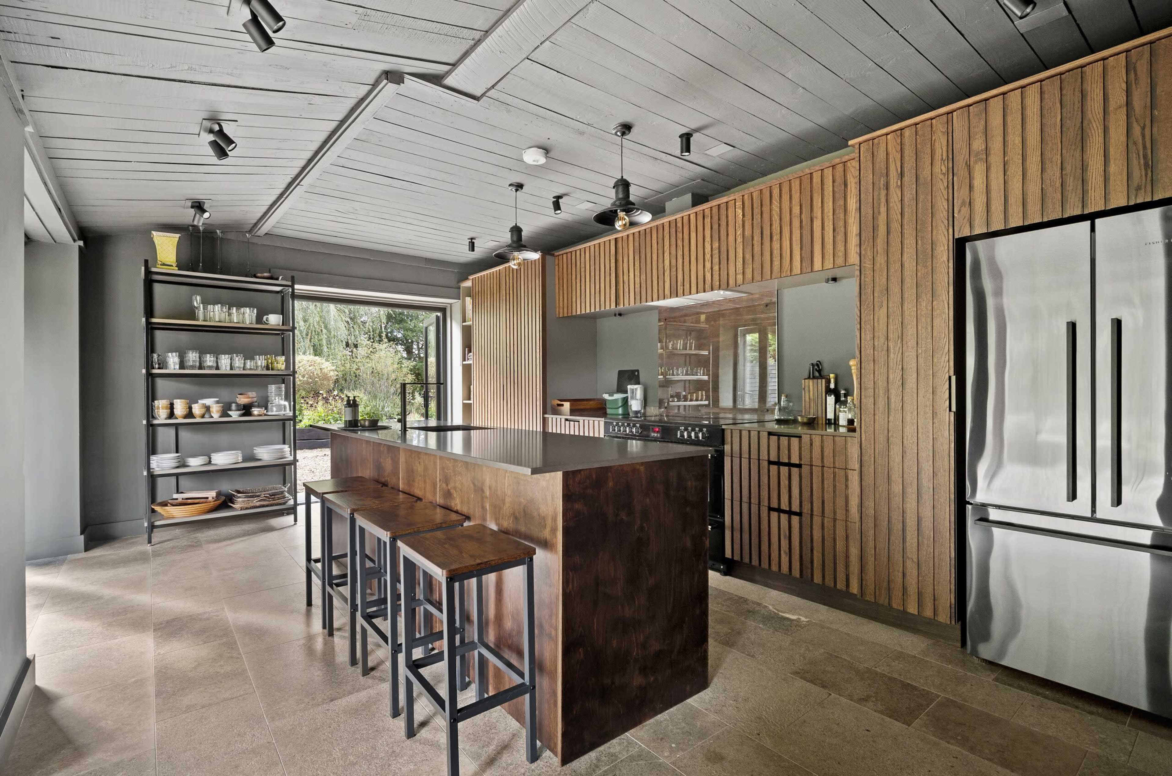 The image shows a modern kitchen with a wood island, metal bar stools, open shelving, and large windows that provide natural light.