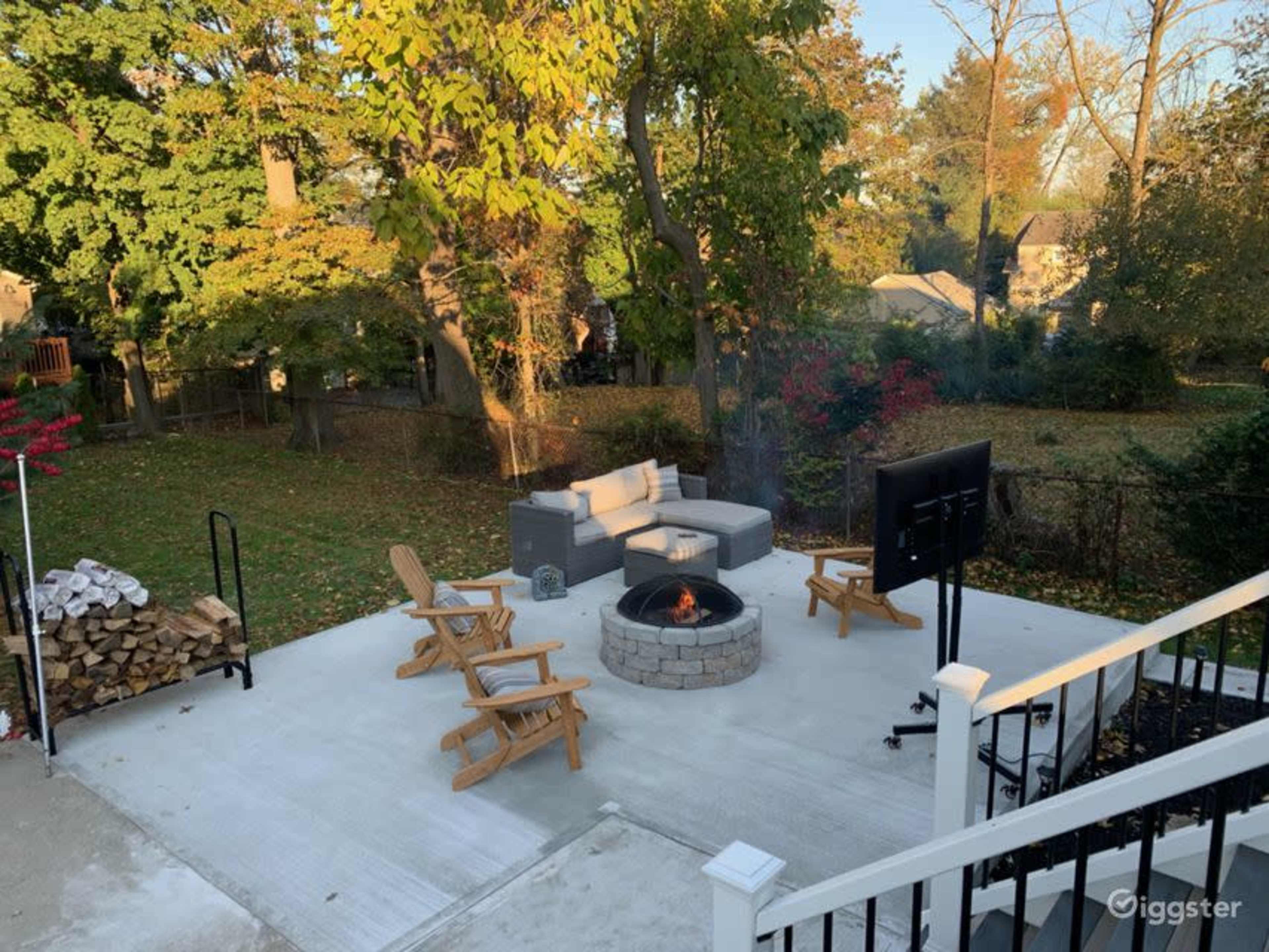 A backyard patio with a fire pit surrounded by cushioned seating and wooden chairs, framed by trees and a stack of firewood.