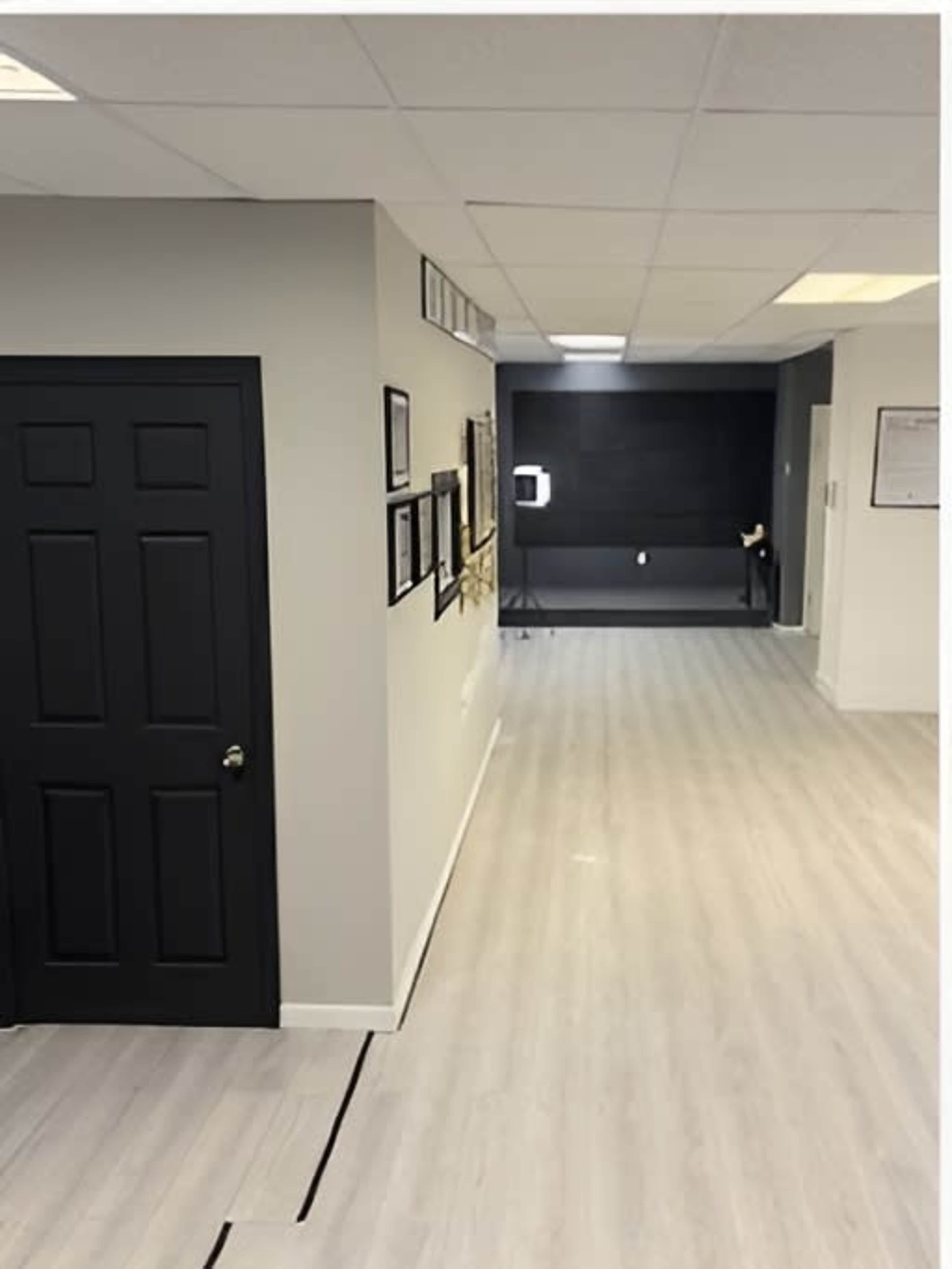 The image shows a well-lit hallway with light wood flooring, featuring a black door on the left and framed pictures on the gray walls.