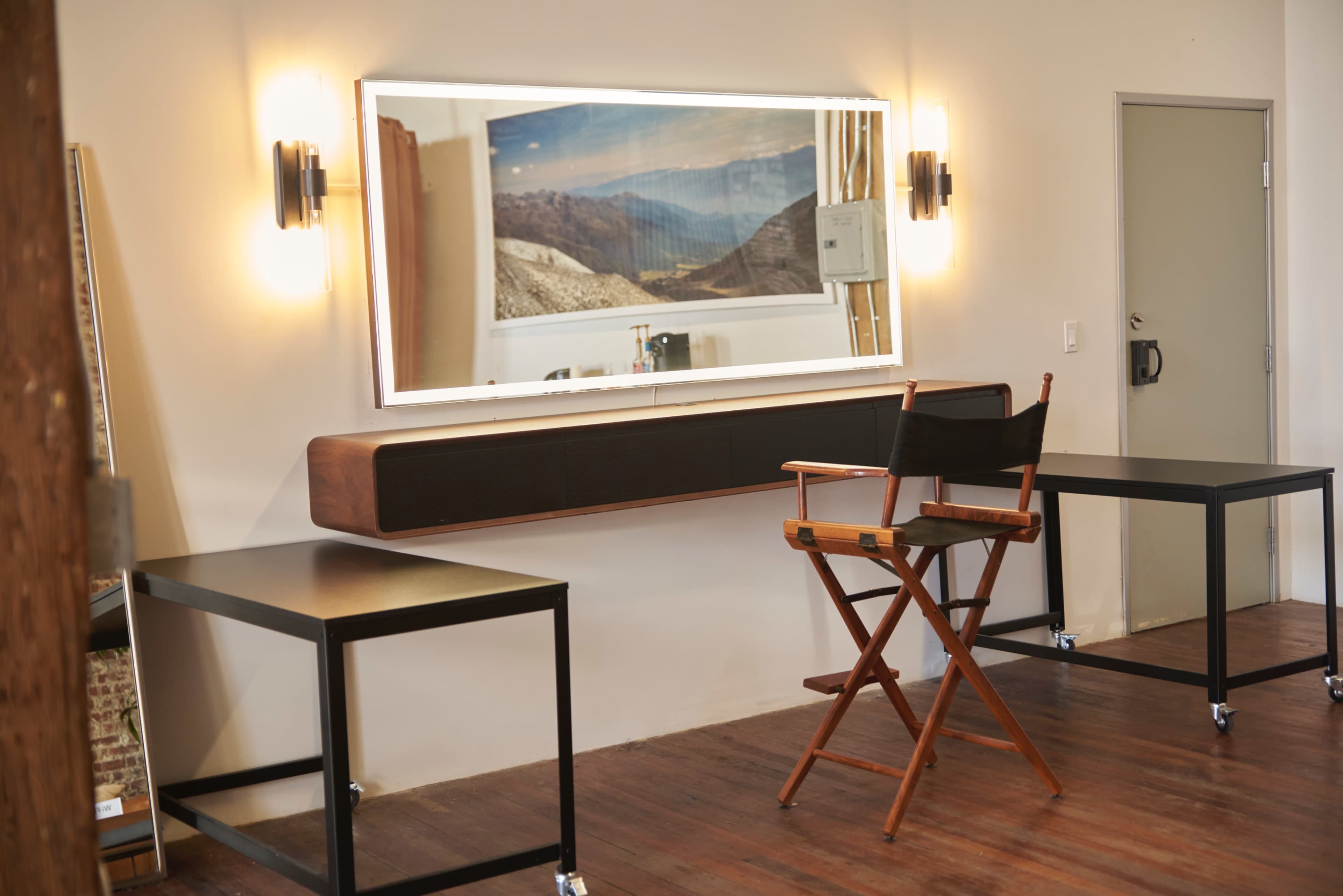 The image shows a bright room featuring a long mirror above a wooden table, with a director's chair and two black tables on wheels.