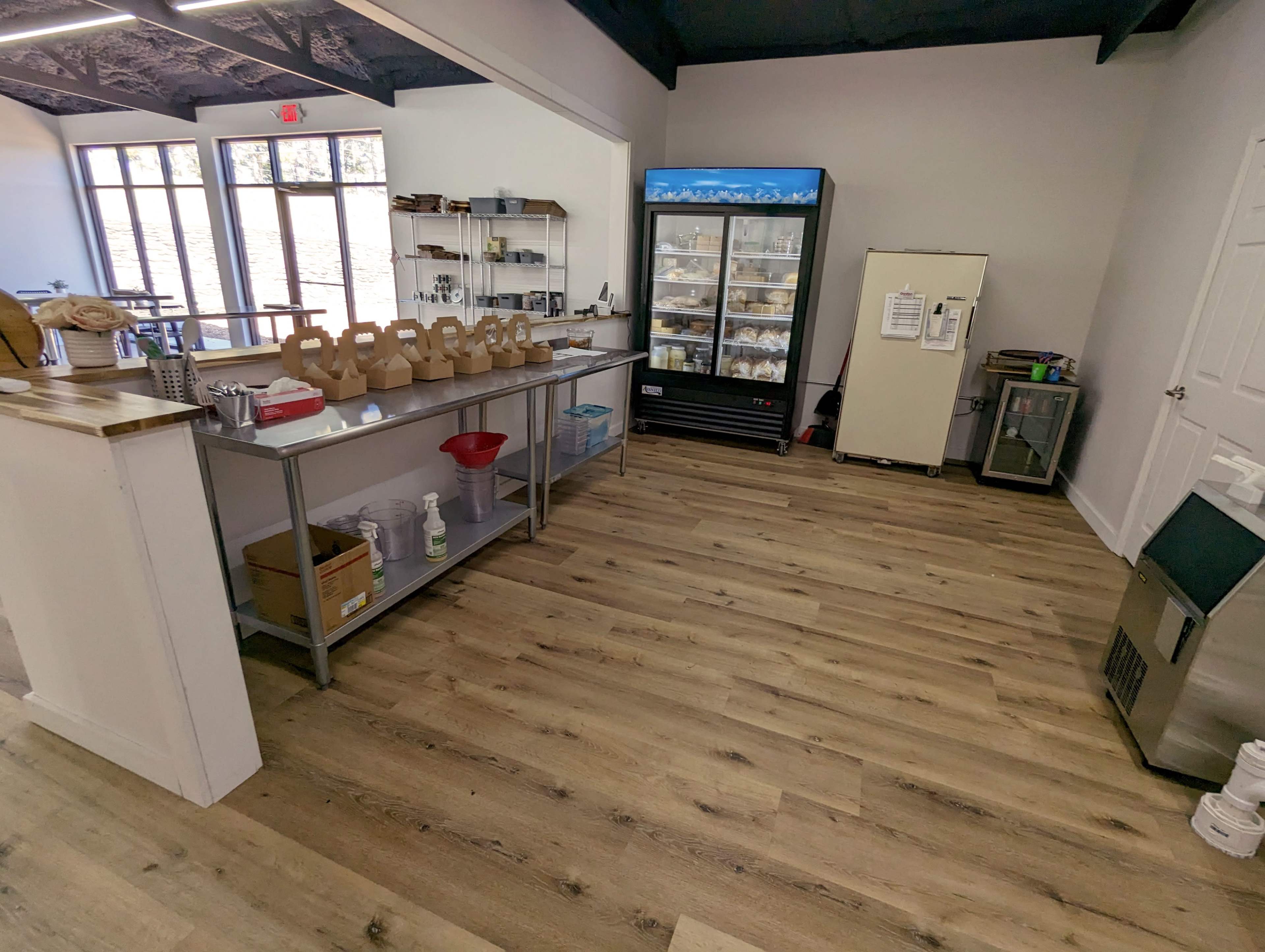 A clean, minimalistic interior of a store or café, featuring a service counter with various items, a glass display fridge, and a beige refrigerator against the wall.