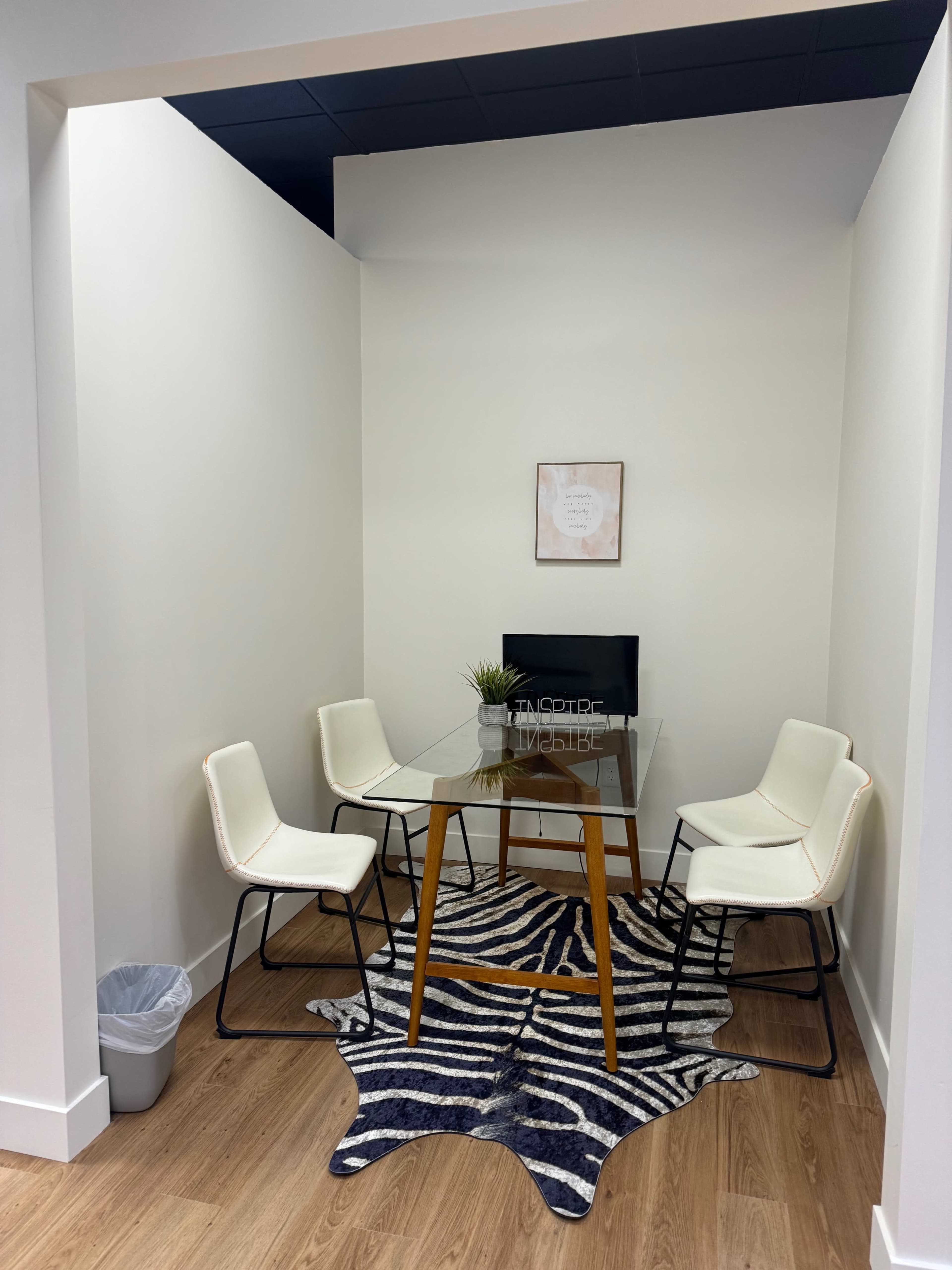 A small meeting room features a glass table surrounded by four white chairs on a zebra-striped rug, with a potted plant and a piece of wall art in the background.