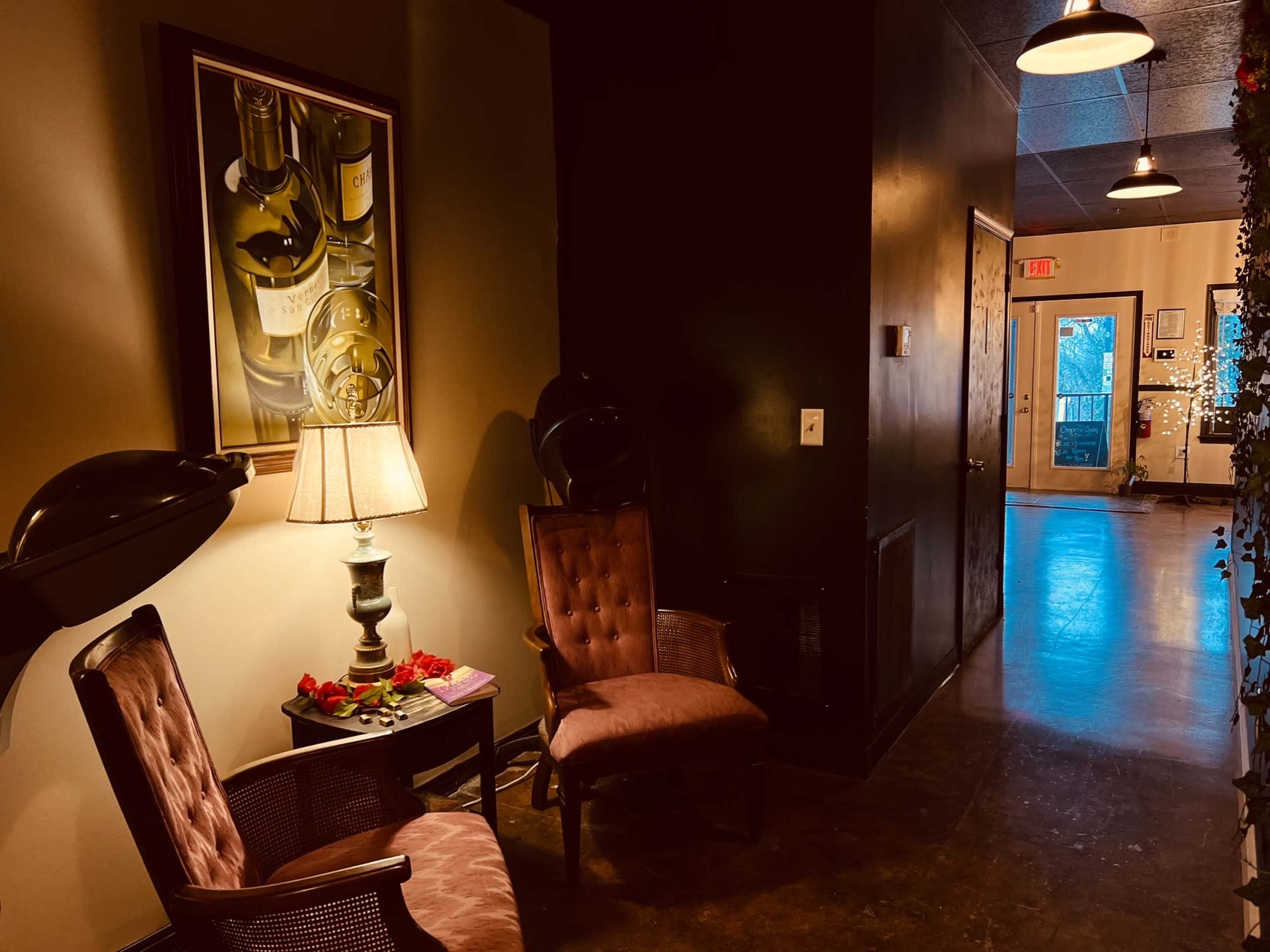 The image shows a dimly lit hallway with two chairs placed beside a lamp, and a door at the end leading outside, flanked by decorative elements.