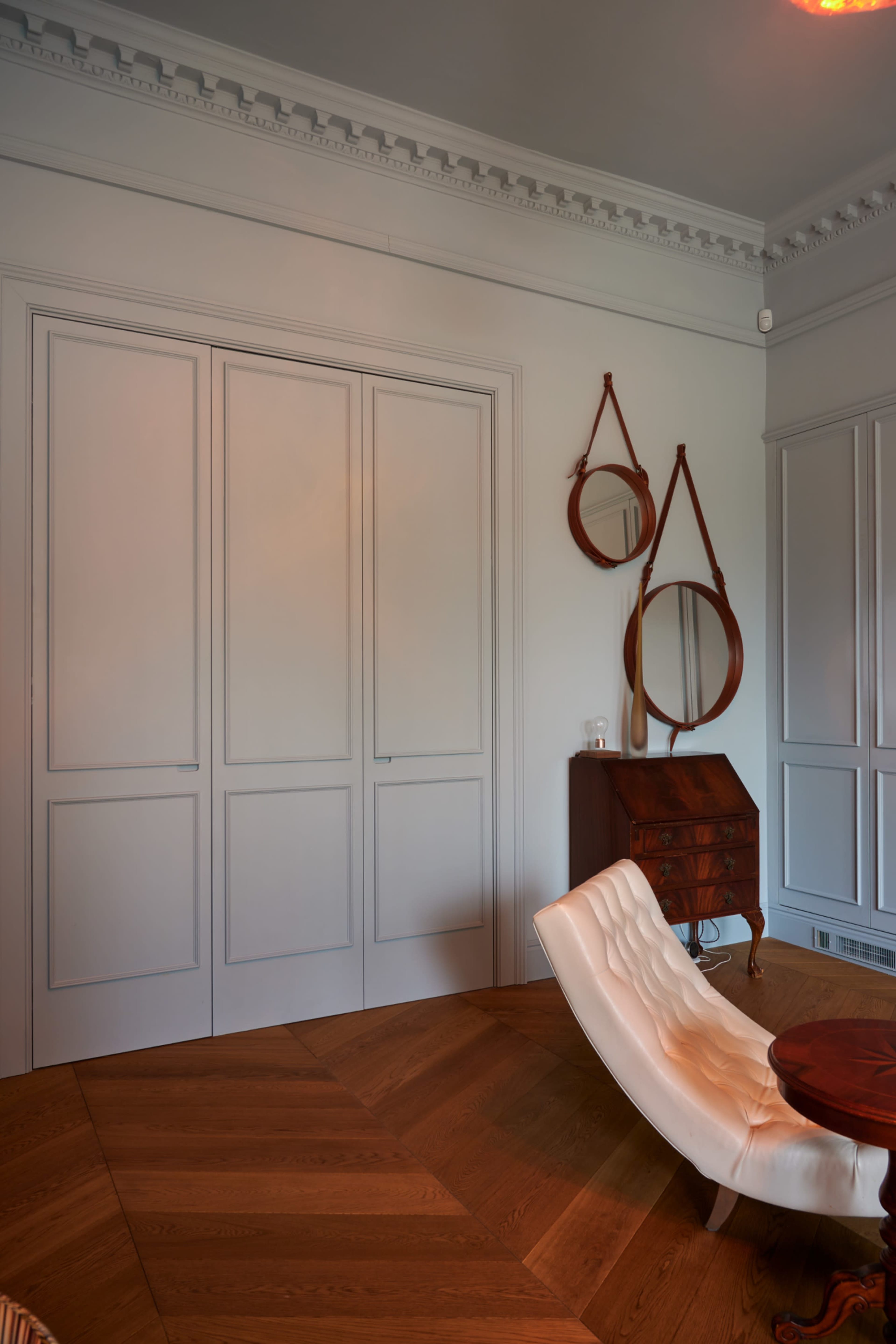 A room with light-colored walls, a wooden floor arranged in herringbone pattern, a small desk, and two round mirrors mounted on the wall.