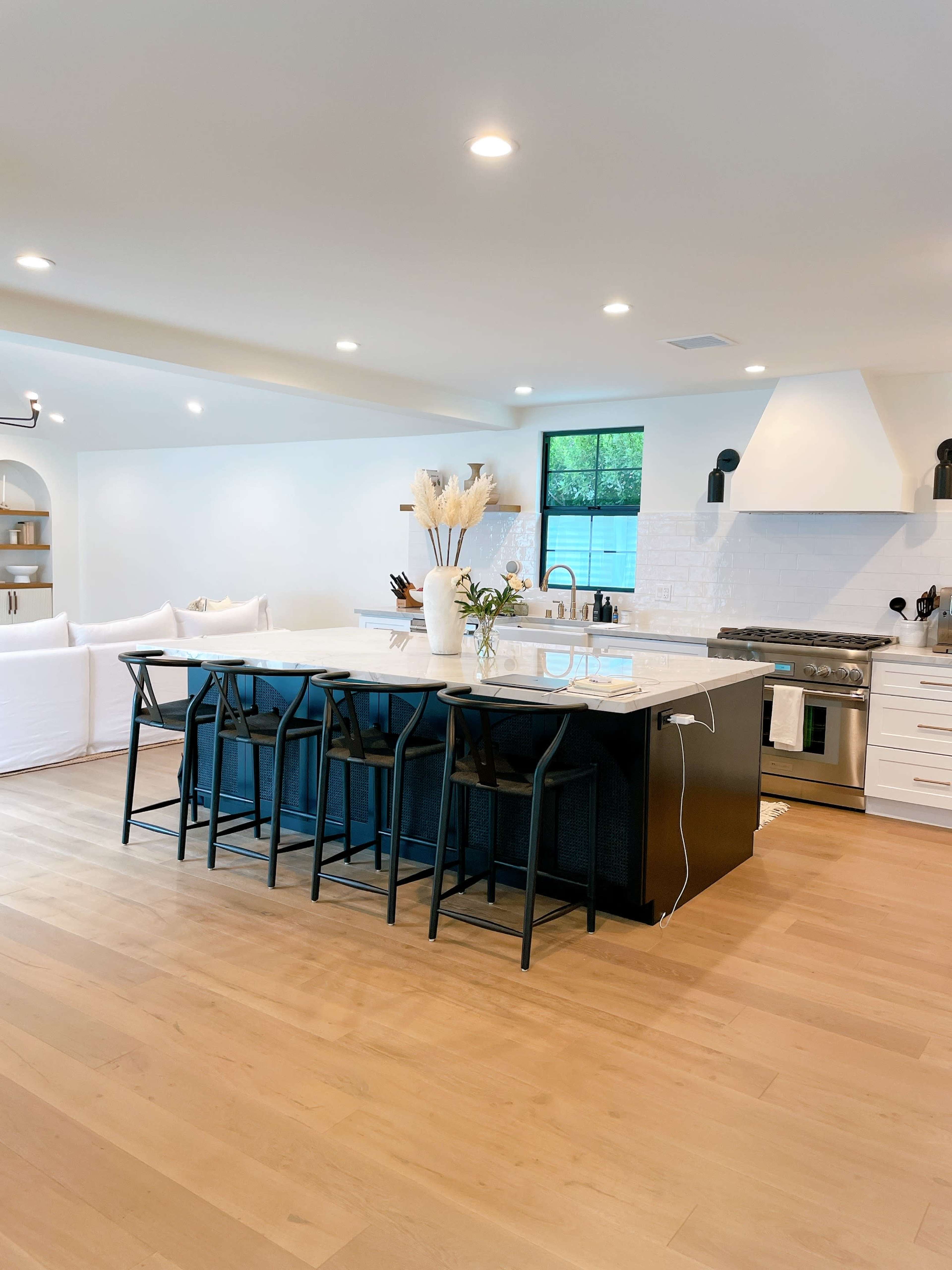 A modern kitchen features a large island with black chairs, white cabinets, and a stainless steel stove against a light background.