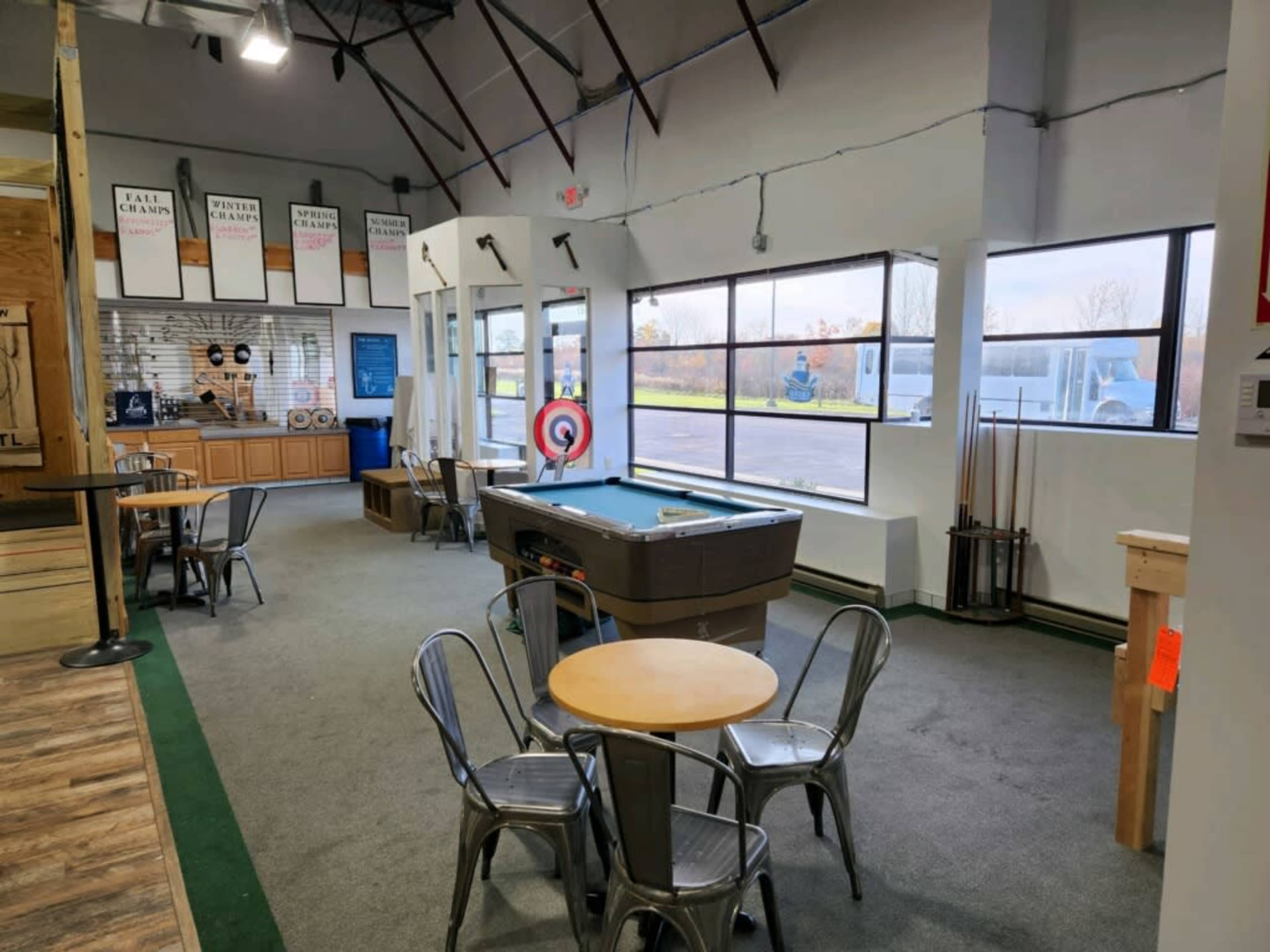 A spacious indoor recreational area with a target setup, a pool table, and several metal chairs arranged around small tables.