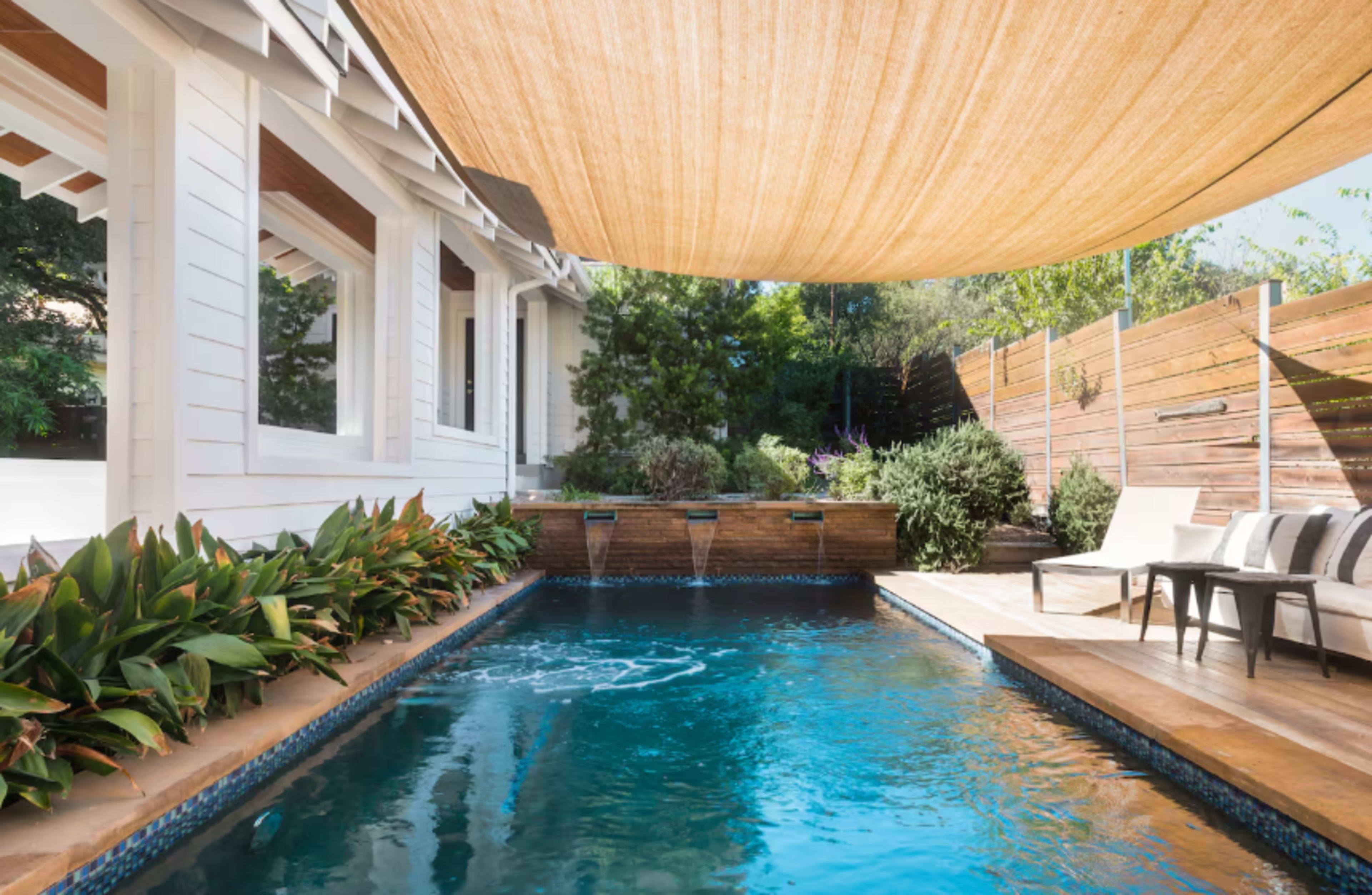 The image shows a swimming pool surrounded by lush greenery and a wooden deck with lounge chairs, under a large shade sail.