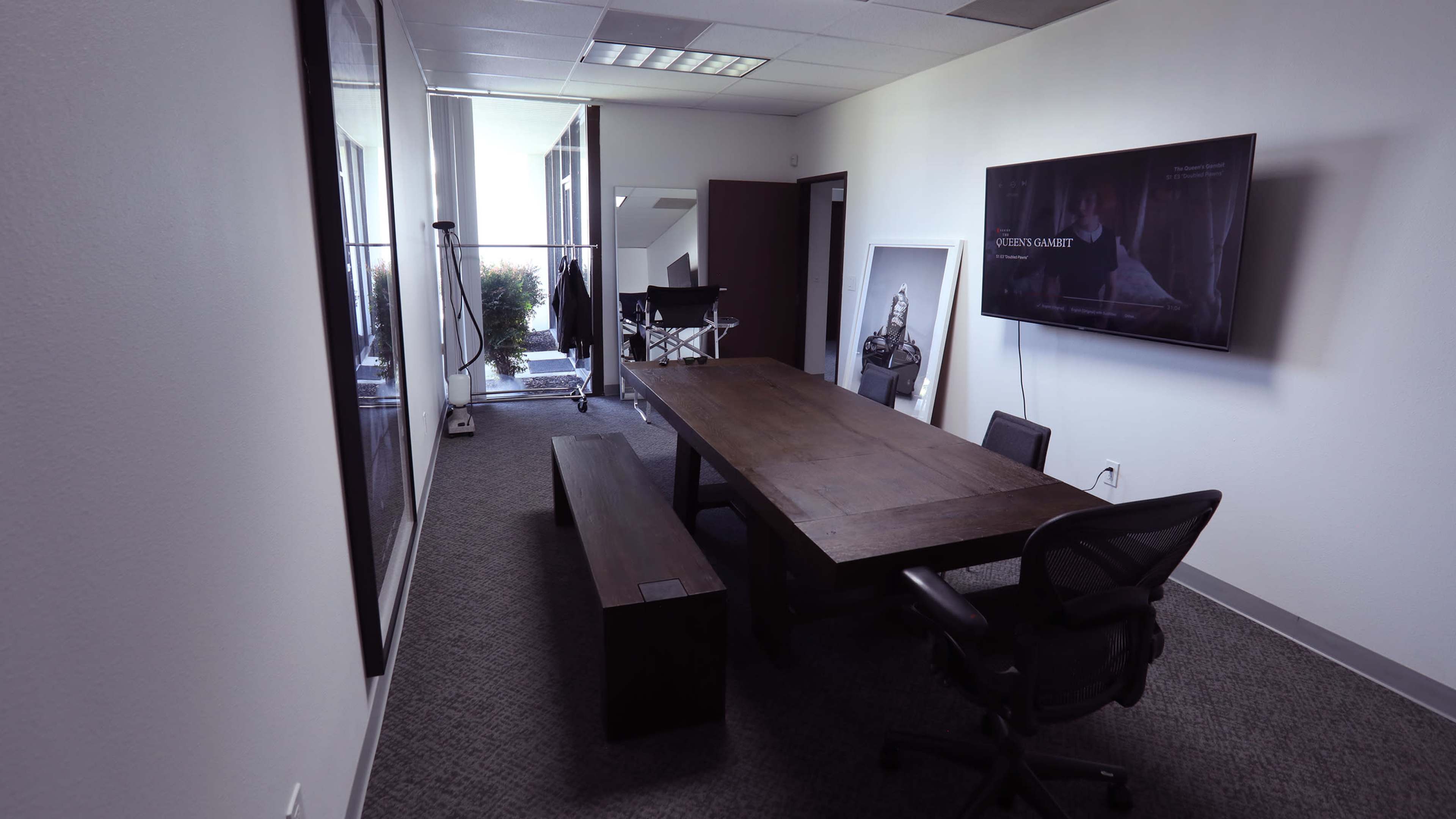A modern conference room with a large wooden table, black chairs, a television mounted on the wall, and natural light filtering through a window.