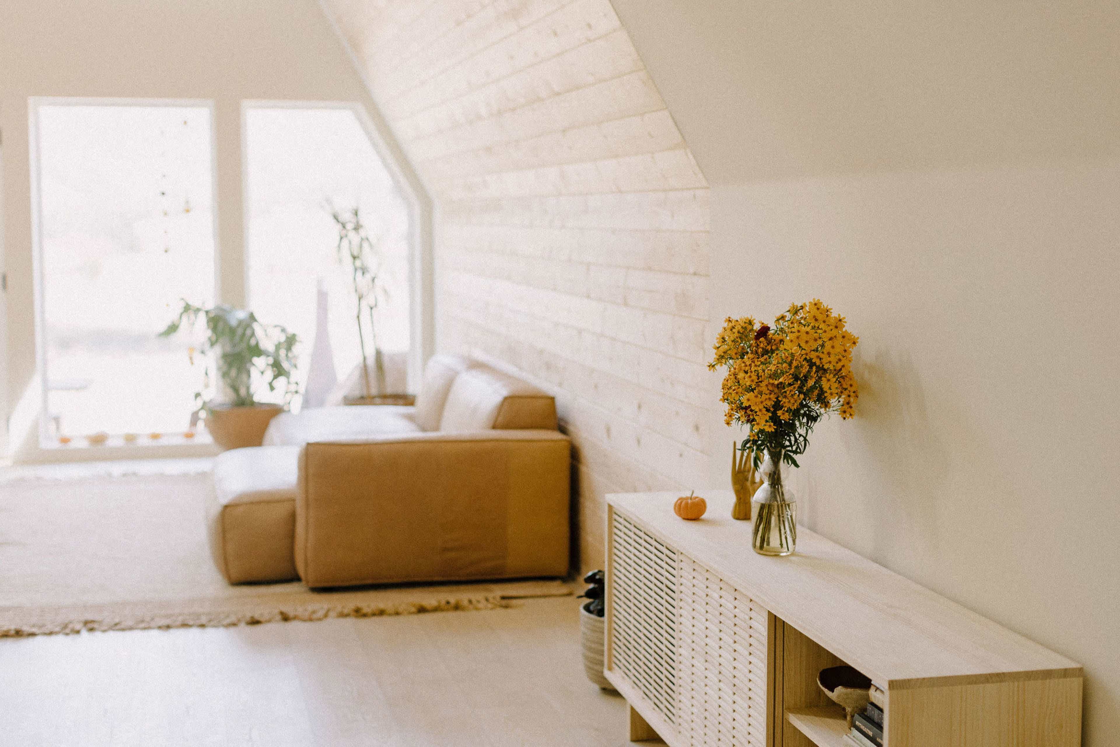 A spacious living room features a light-colored wooden accent wall, a tan couch, a vase of yellow flowers on a low cabinet, and large windows with natural light.
