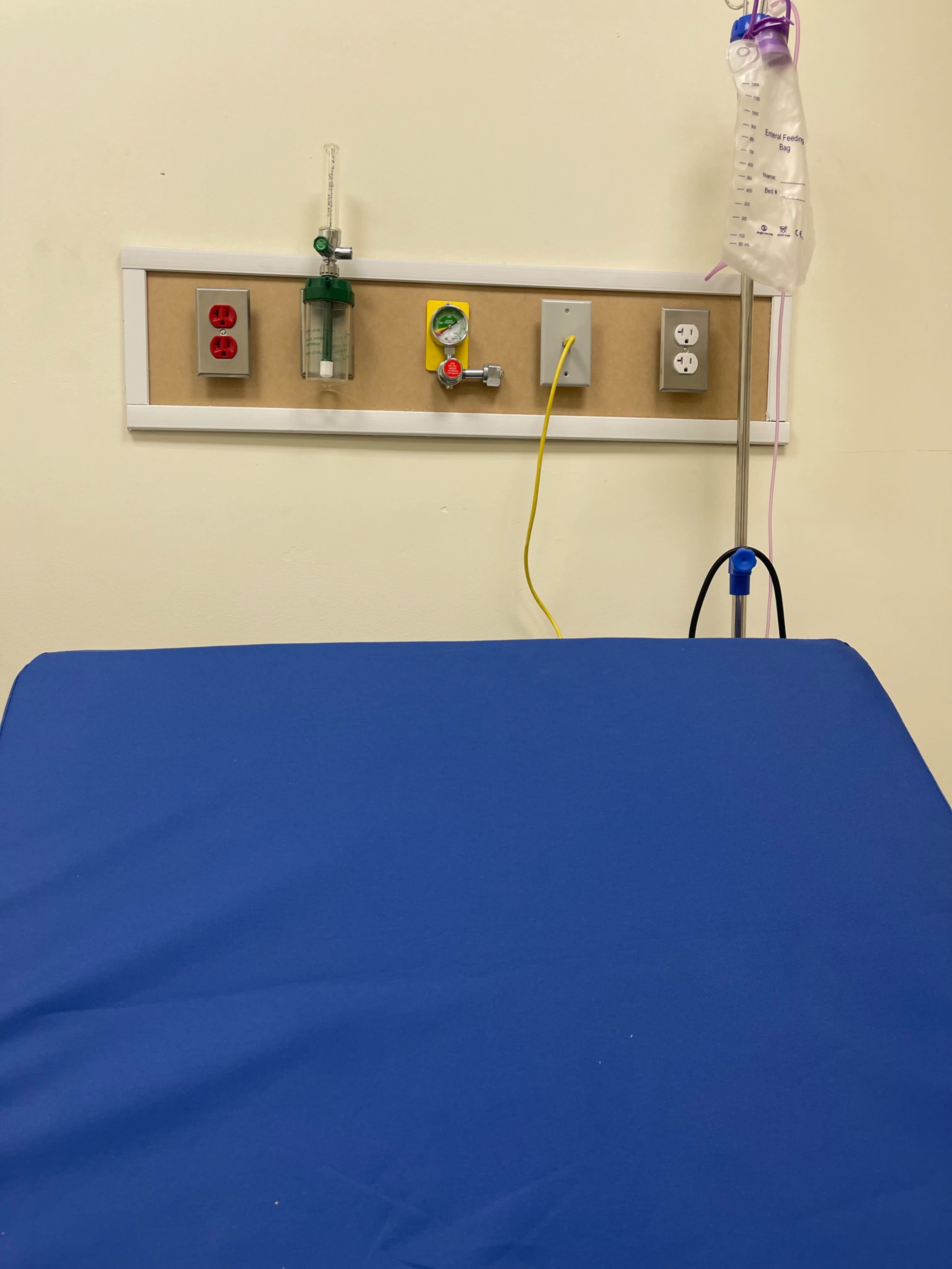 The image shows a hospital bed with a blue sheet and medical equipment, including an IV pole and a wall-mounted panel featuring electrical outlets and a faucet.