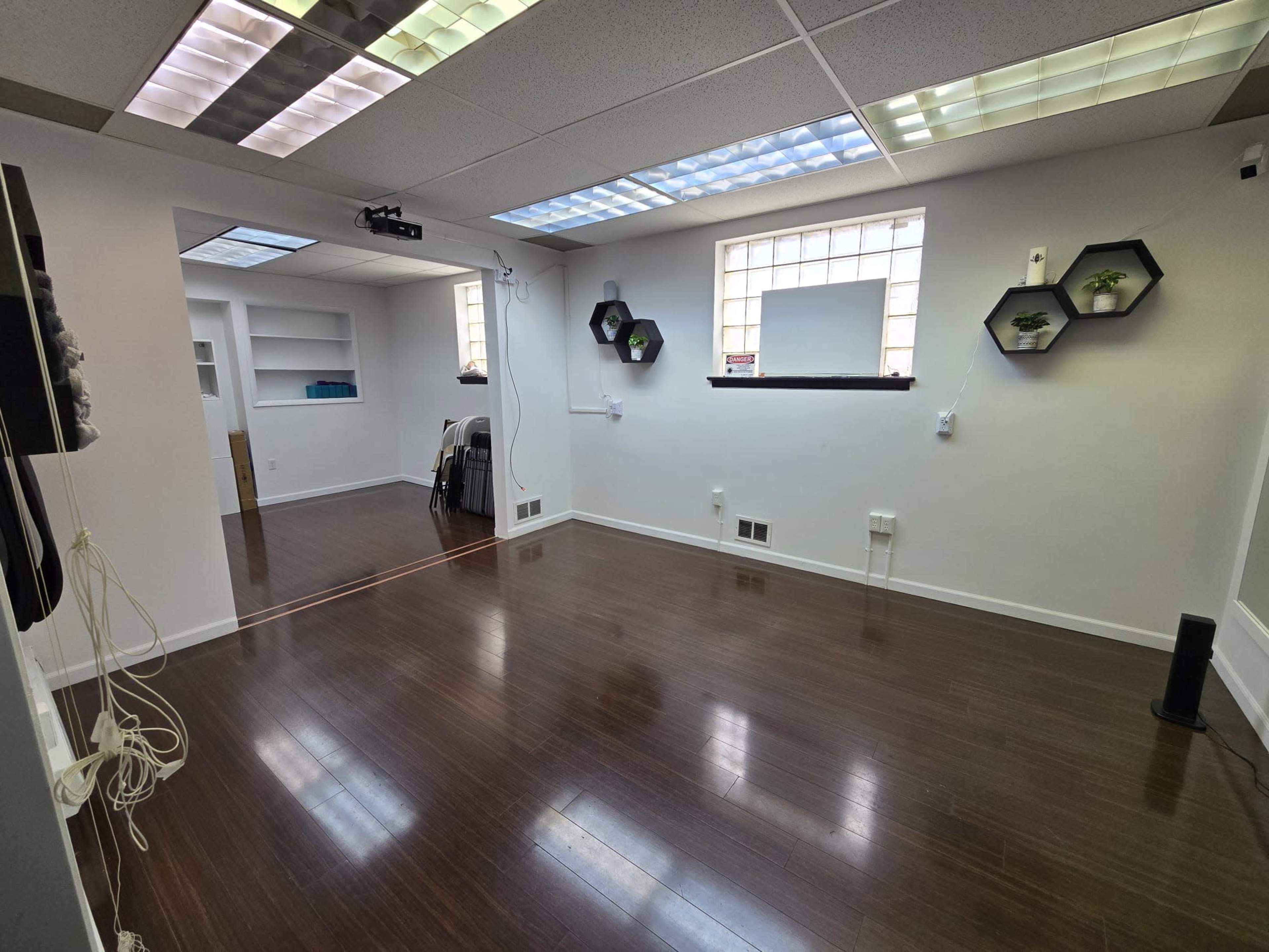 A spacious, brightly lit room with wooden flooring, white walls, and geometric wall decor, featuring plants in hexagonal fixtures.