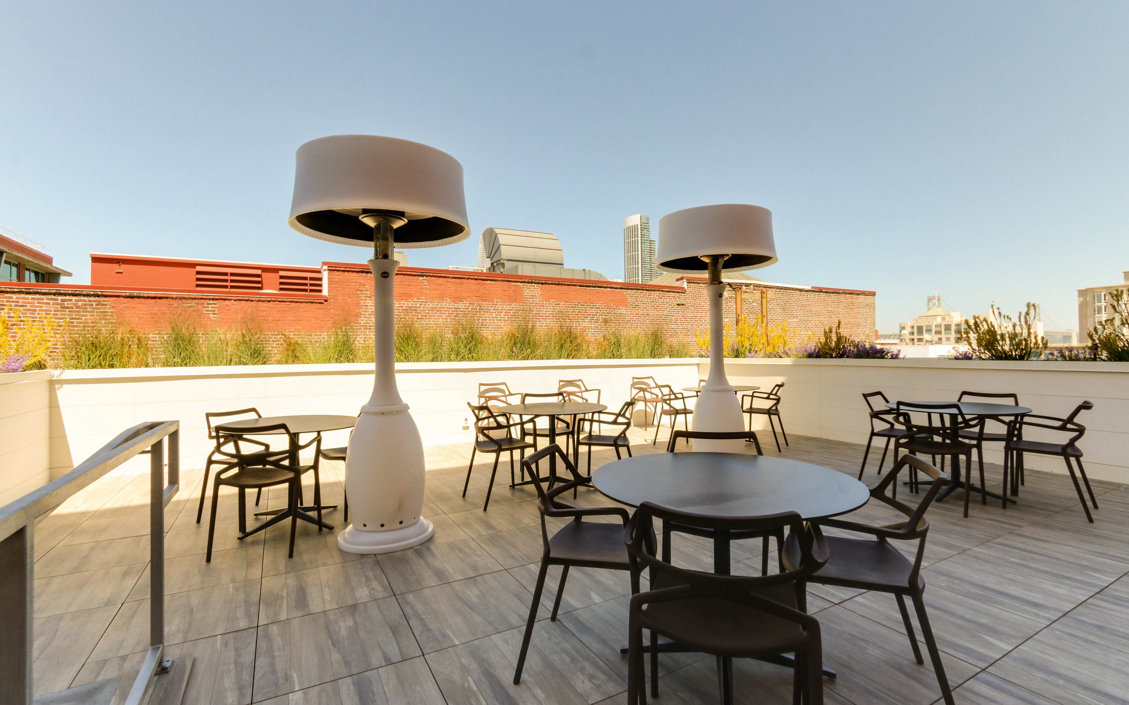 An outdoor seating area with several round tables and chairs, complemented by tall lamps, set against a backdrop of urban buildings.