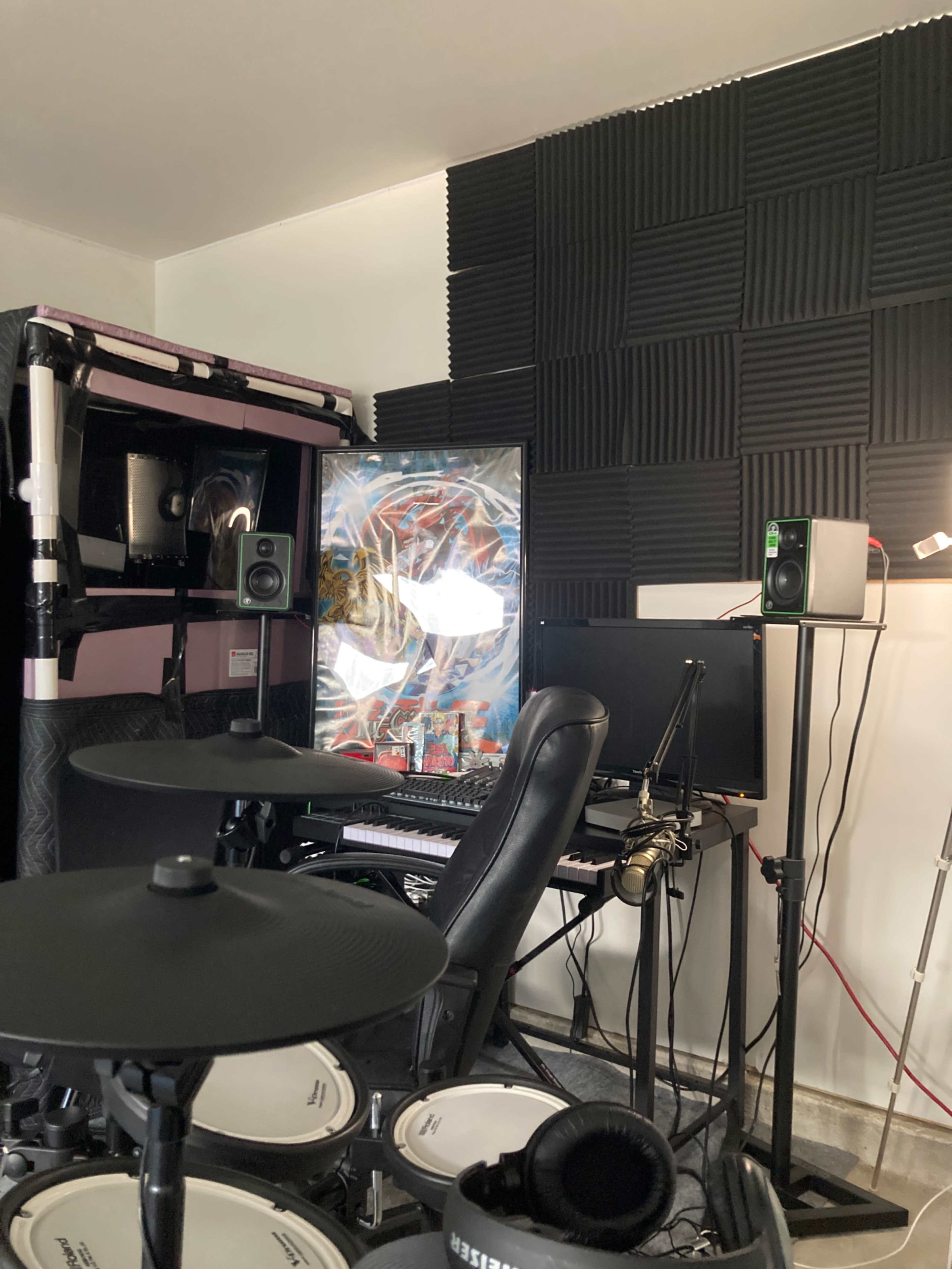A music production space features a drum set, computer setup with a monitor, and acoustic panels on the walls.