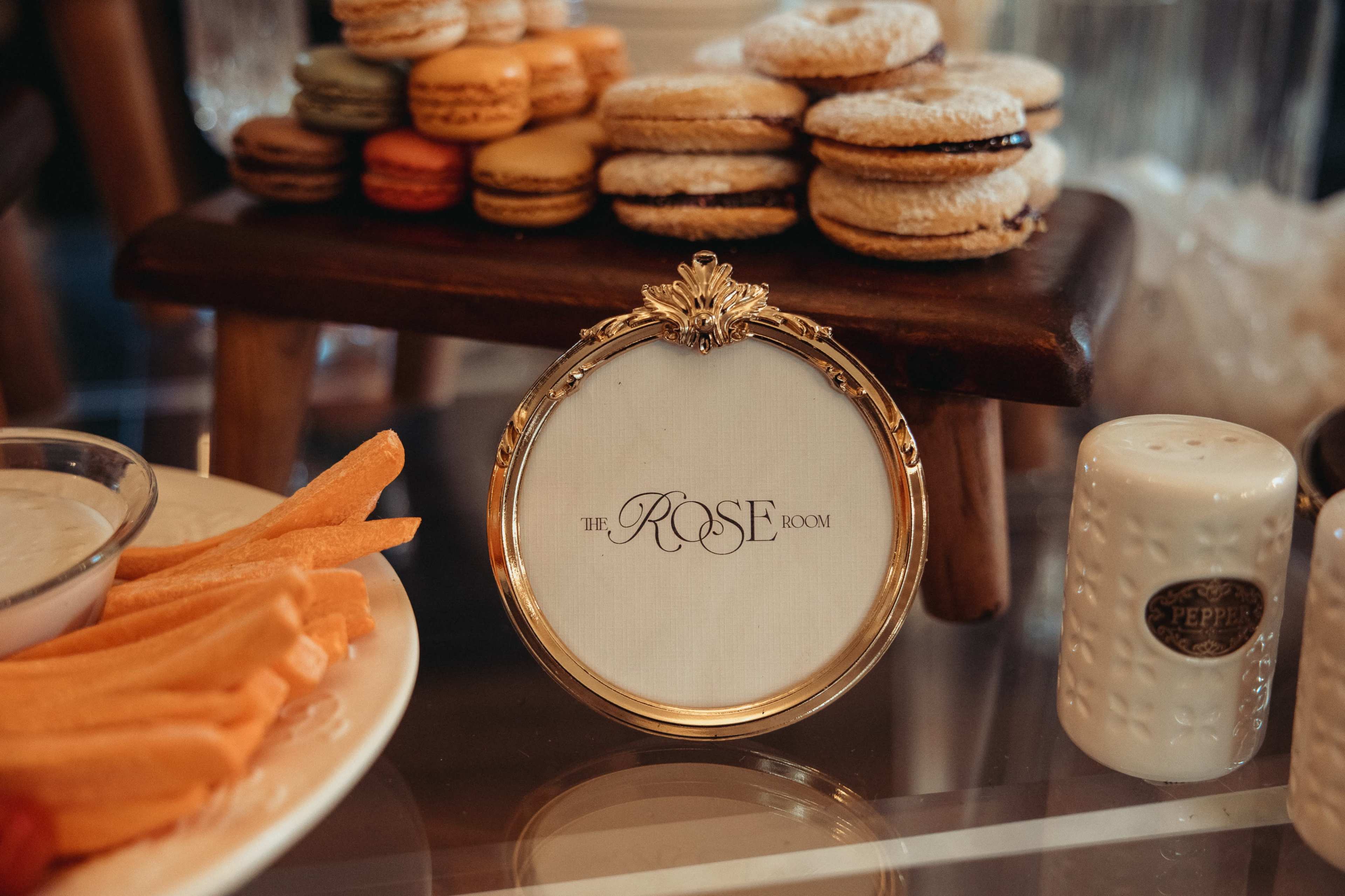 A decorative sign reading "The Rose Room" is placed on a table next to an assortment of colorful macarons and a plate of fresh vegetables.