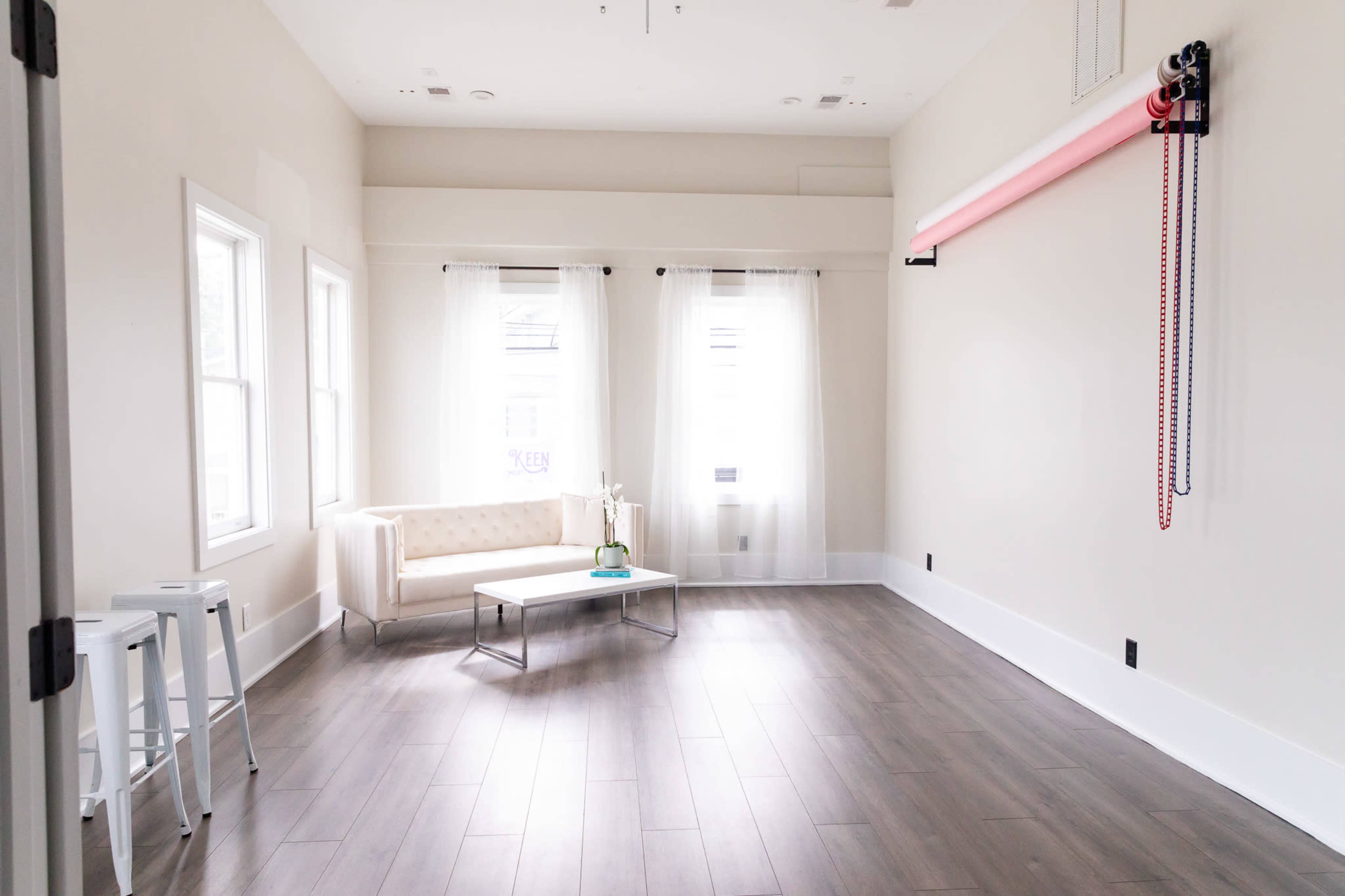 A minimalist room with light wooden floors, a white couch, a coffee table, and large windows with sheer curtains.