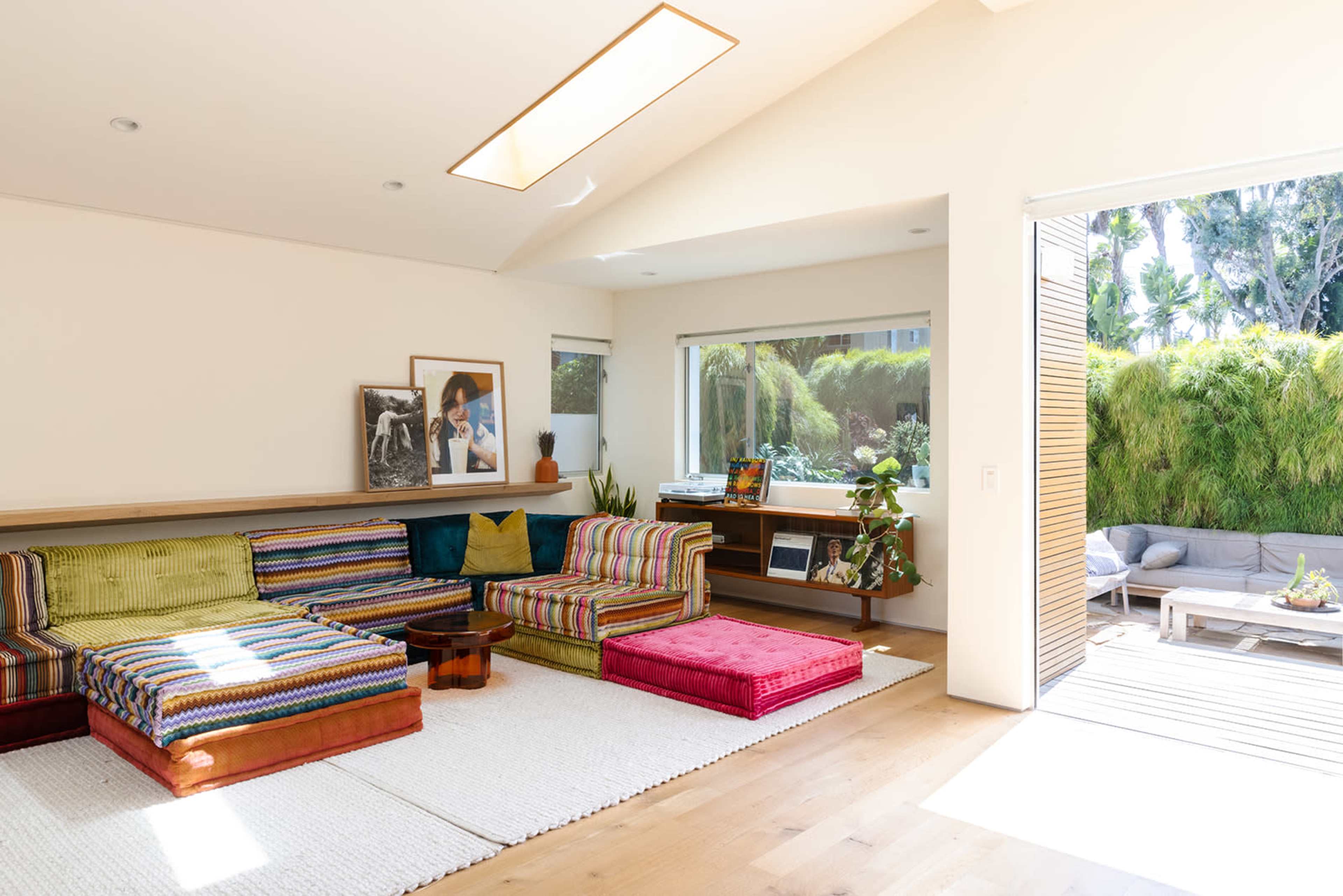 The living room features colorful striped seating, a wooden coffee table, and large windows that open to a green outdoor area.