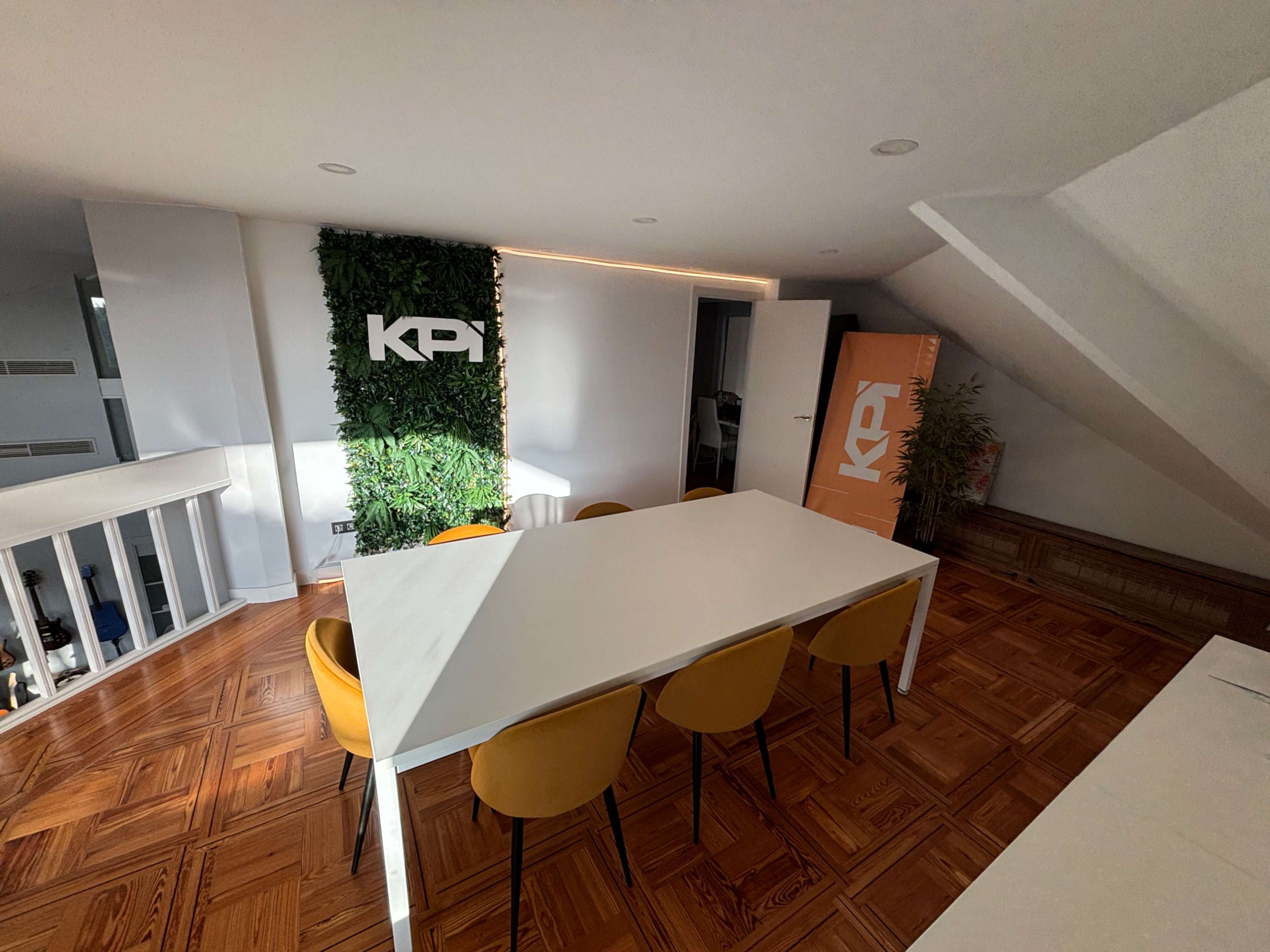 The image shows a well-lit meeting room featuring a large white table surrounded by yellow chairs, with green foliage on the wall and branding elements in orange and white.