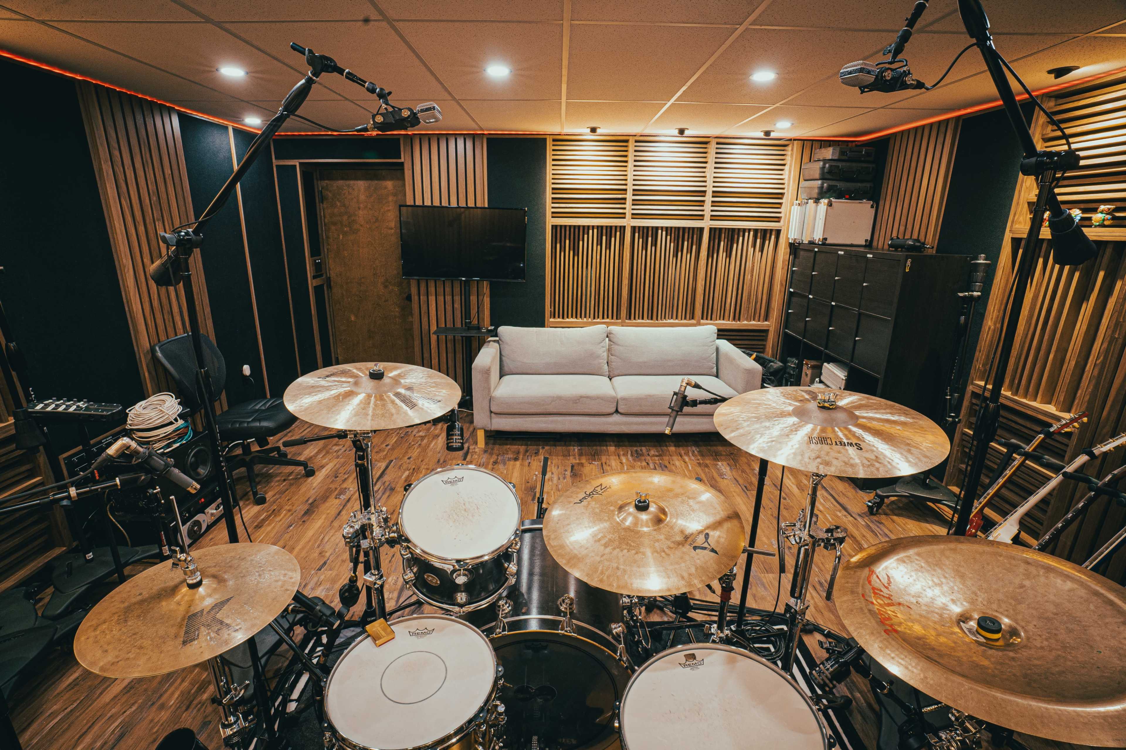 The image shows a well-equipped recording studio with a drum set in the foreground and a cozy seating area in the background.