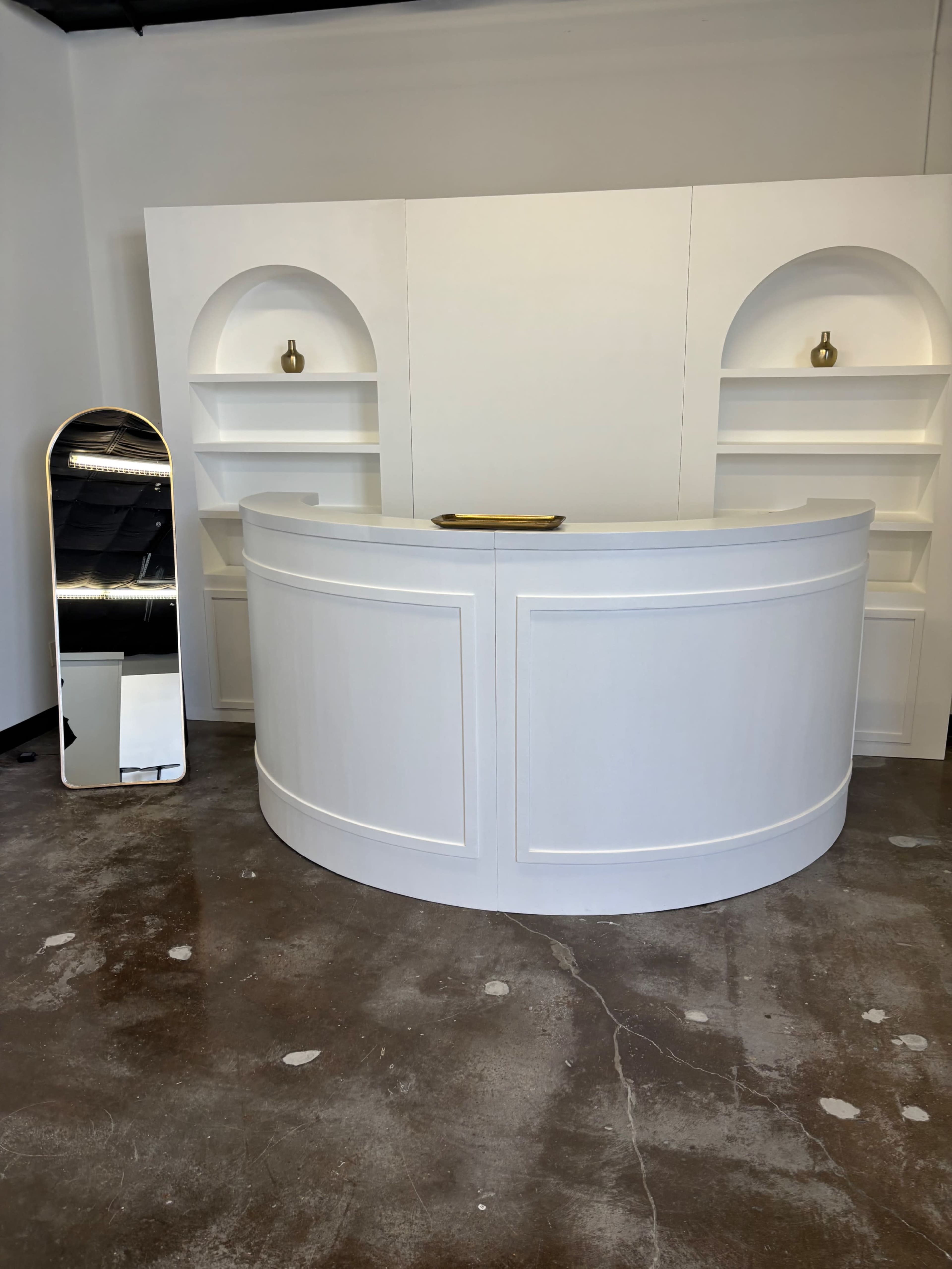 The image shows a curved white reception desk with shelves behind it, accompanied by a tall, mirrored standing mirror on the left, set in a room with a polished concrete floor.