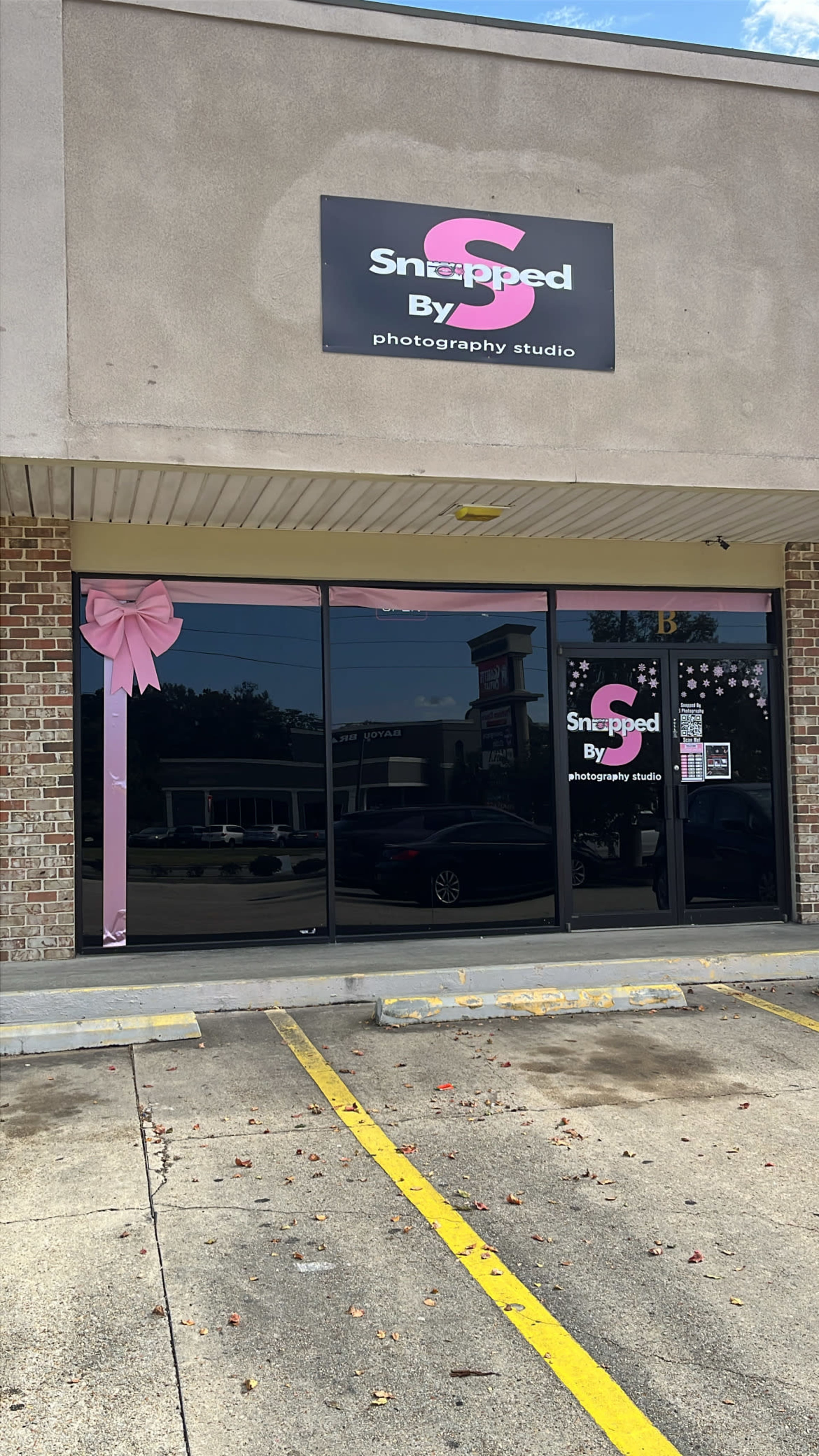 The image shows the exterior of a photography studio named "Snapped By," located in a commercial building with large windows and a pink bow decoration.