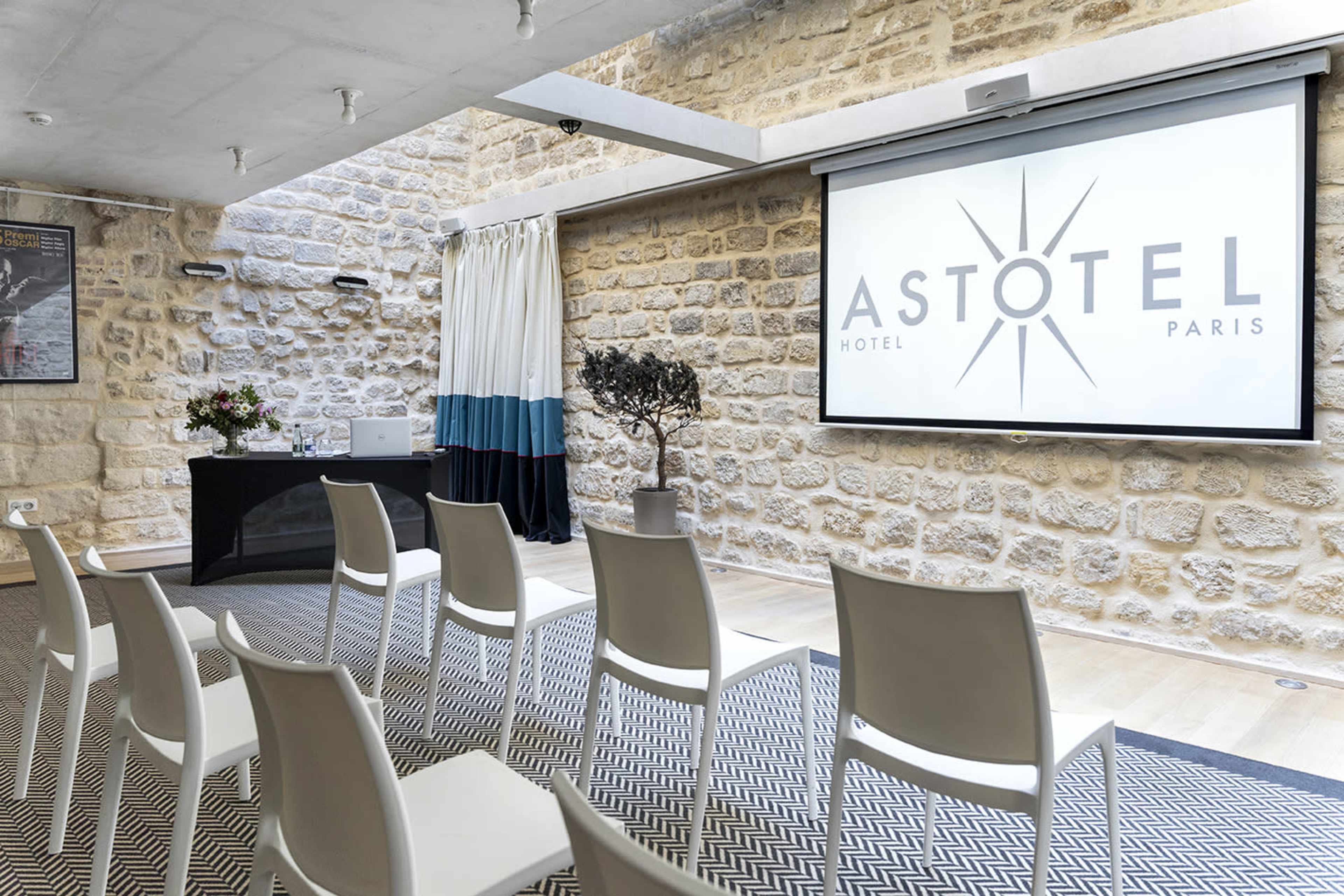 A meeting room with white chairs arranged in rows, a presentation screen displaying the "ASTOTEL PARIS" logo, and stone walls decorated with a potted plant and floral arrangements.