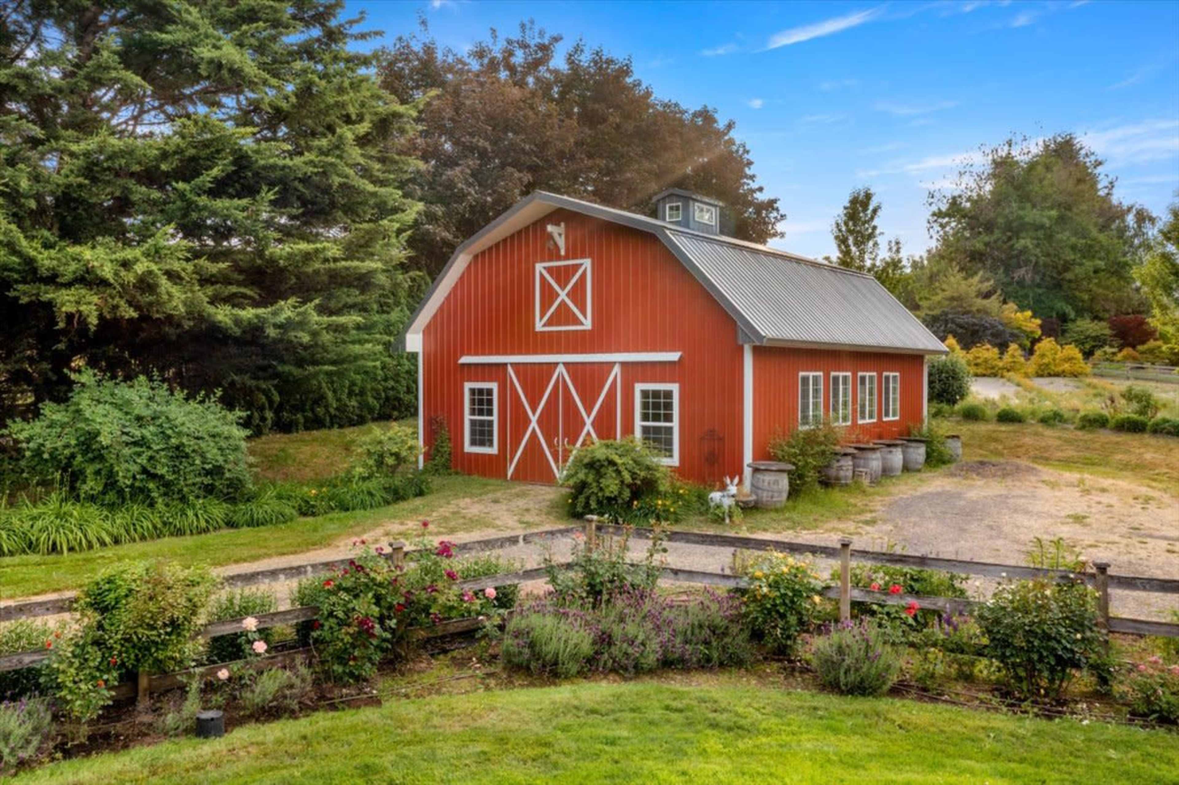A red barn with white trim situated among lush greenery and flowerbeds in a rural setting.