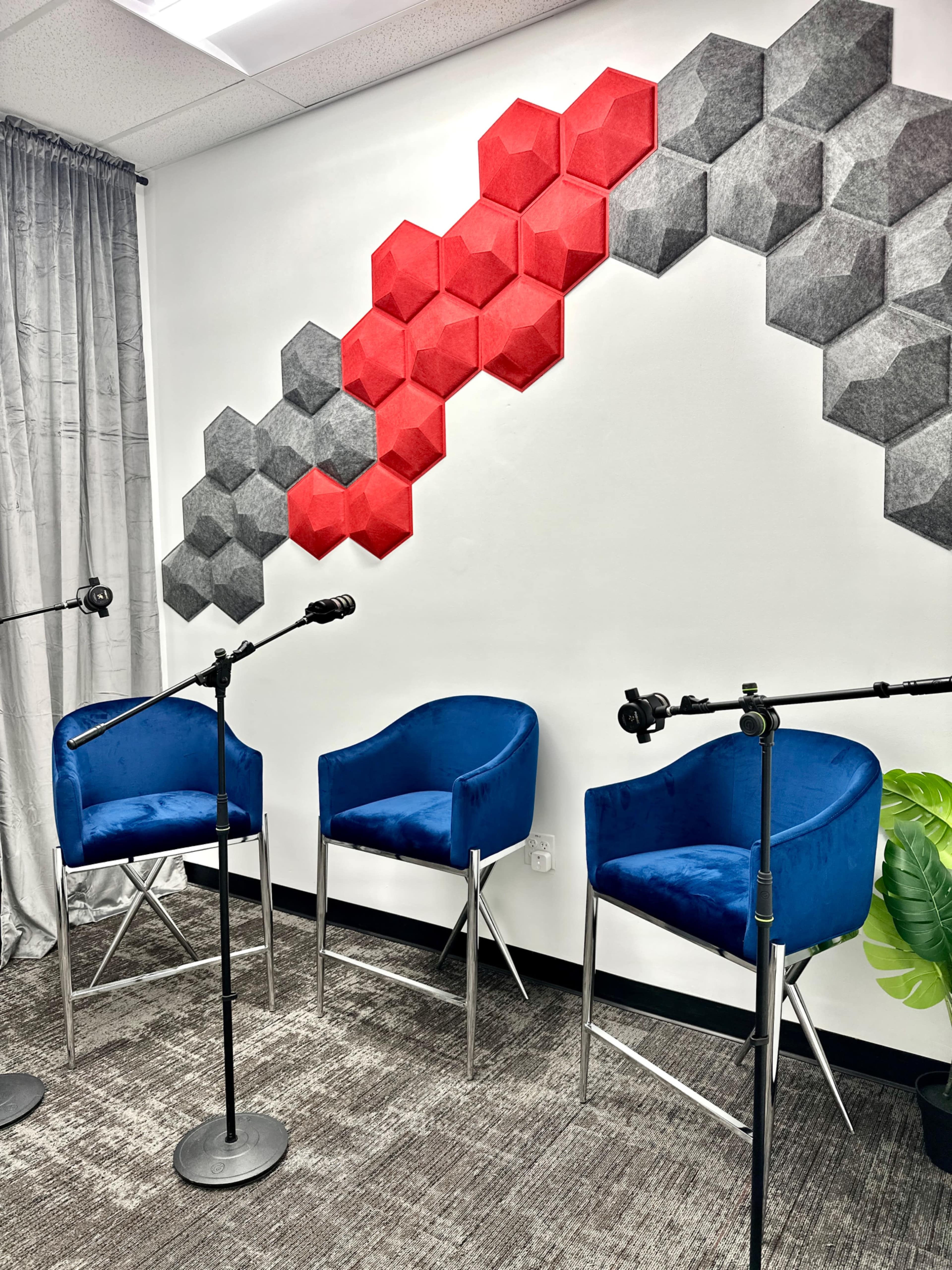 A modern seating area features two blue chairs and microphone stands against a wall adorned with red and gray hexagonal sound panels.