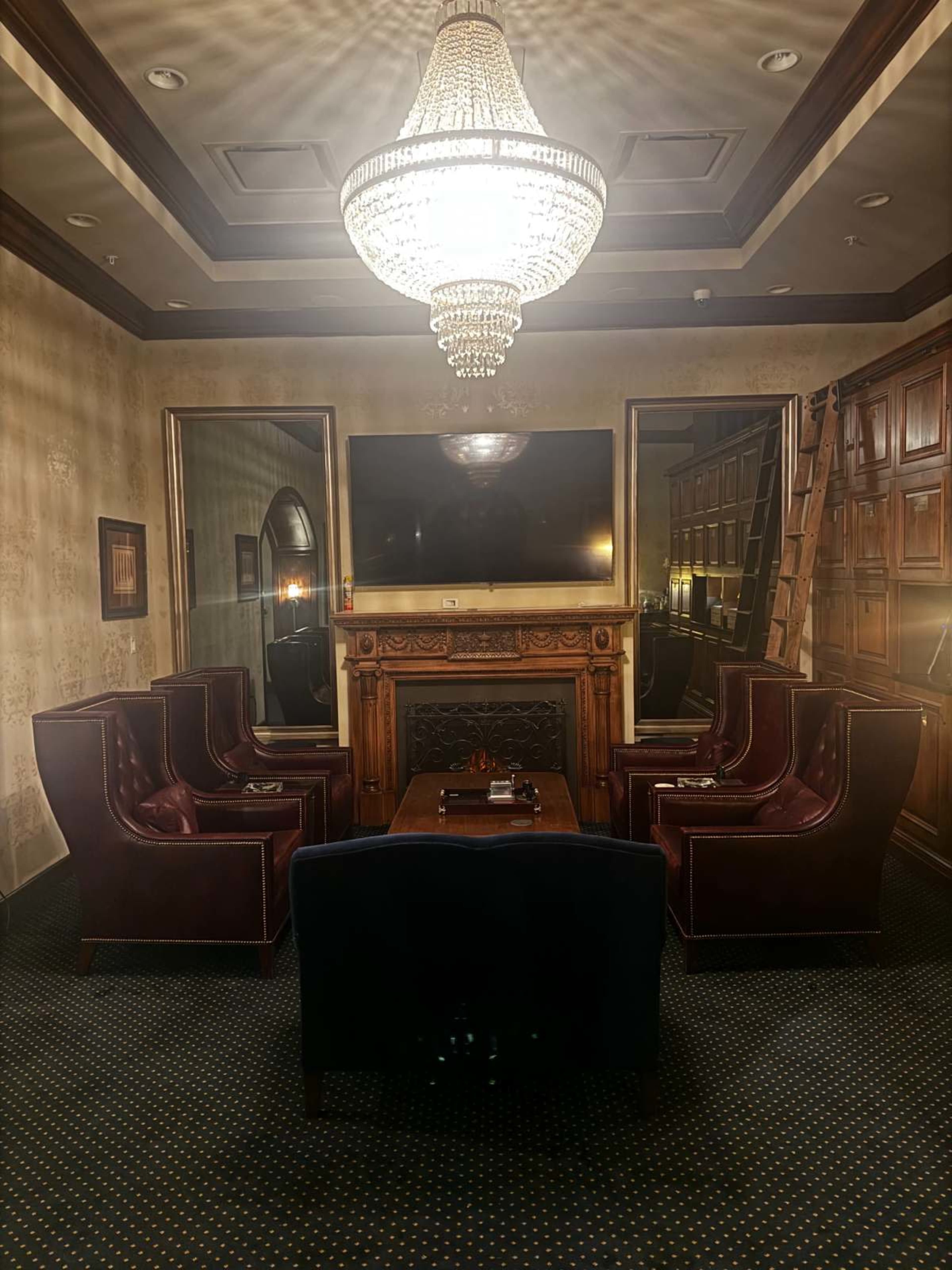 A luxurious lounge features plush, dark leather seating arranged around a central table, with an ornate chandelier overhead and a large screen mounted on the wall.