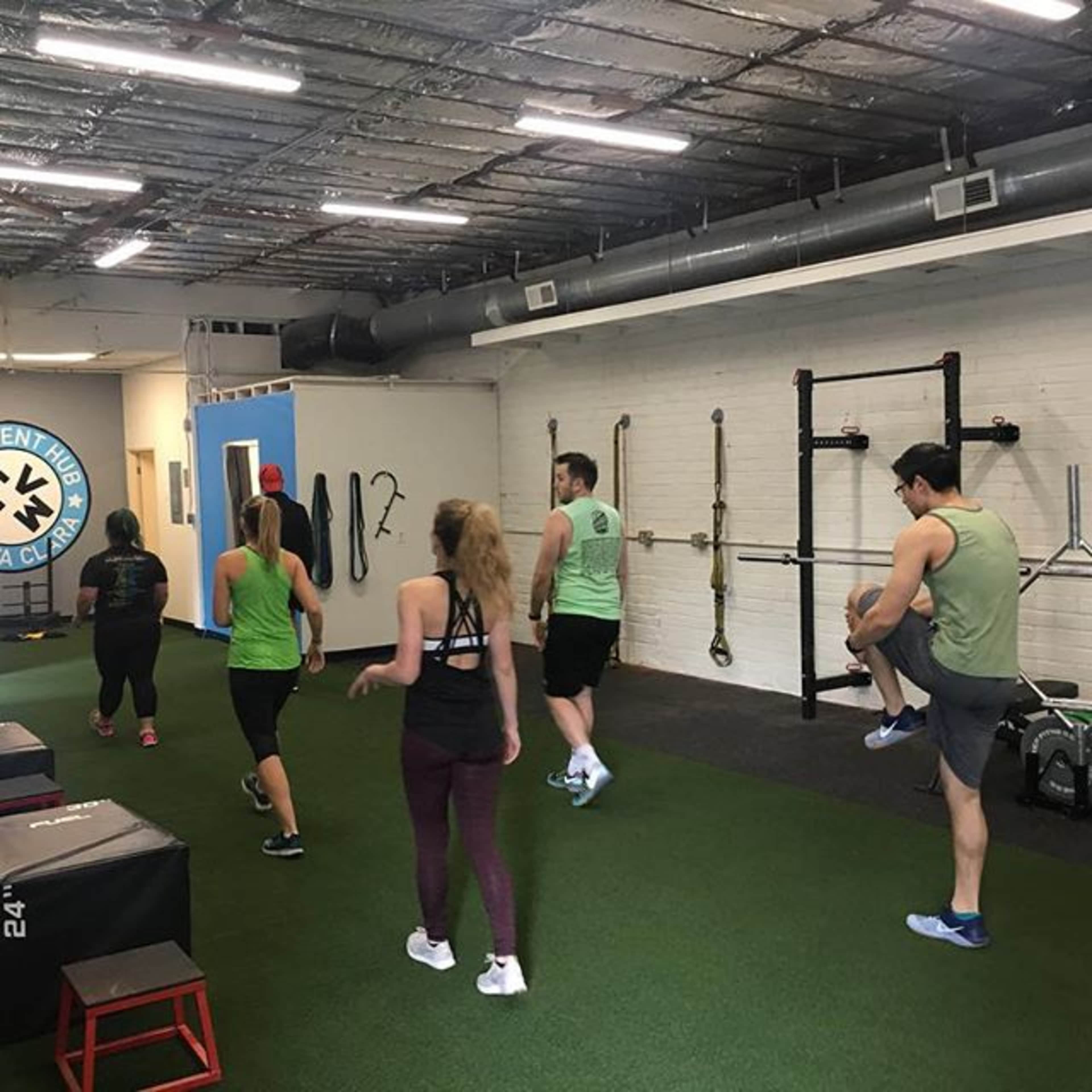 A group of five people is exercising in a gym with green turf flooring and various workout equipment.