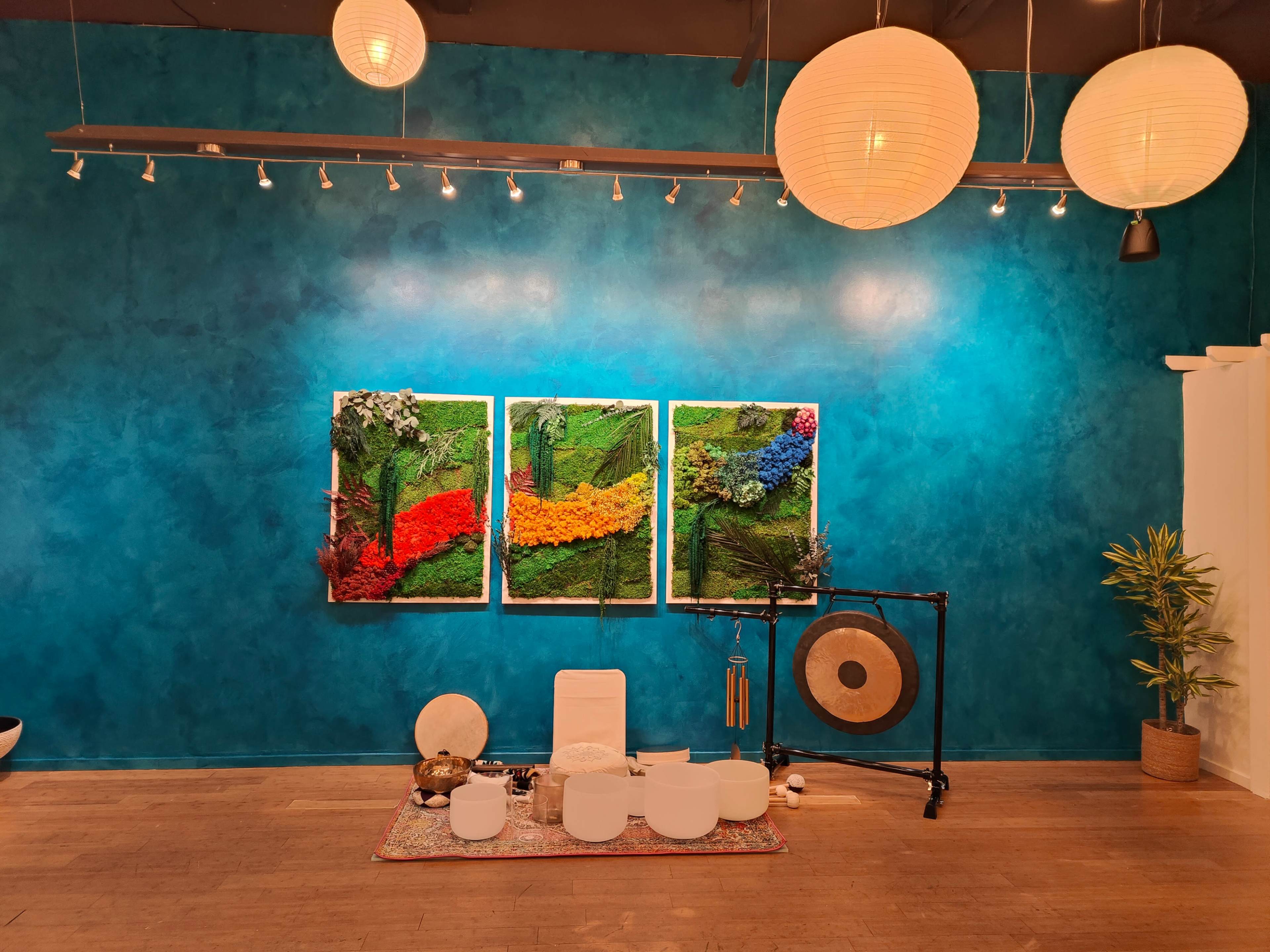 The image shows a vibrant blue wall with three framed artworks made of moss and colorful plants above a serene seating area featuring white furniture and a gong.