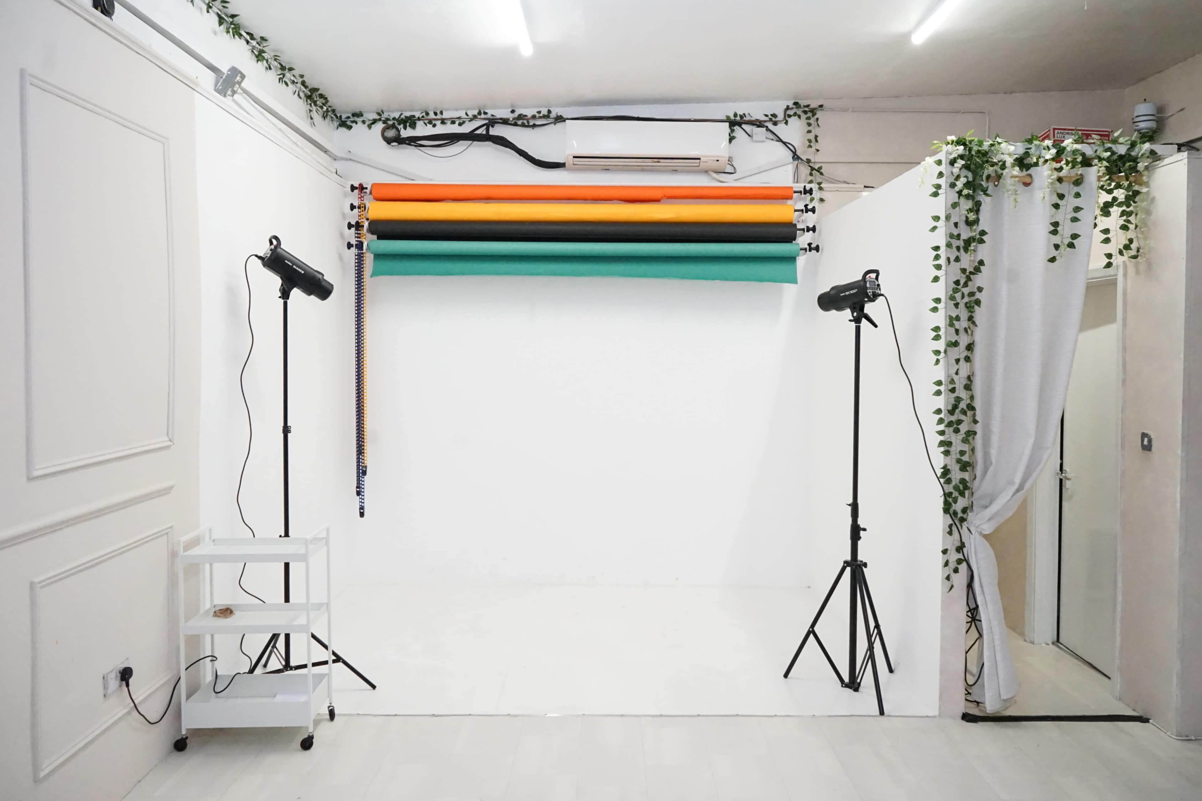 The image shows a photography studio with a white backdrop, two lighting stands, a small cart, and rolls of colored backdrop paper in orange, green, and yellow.
