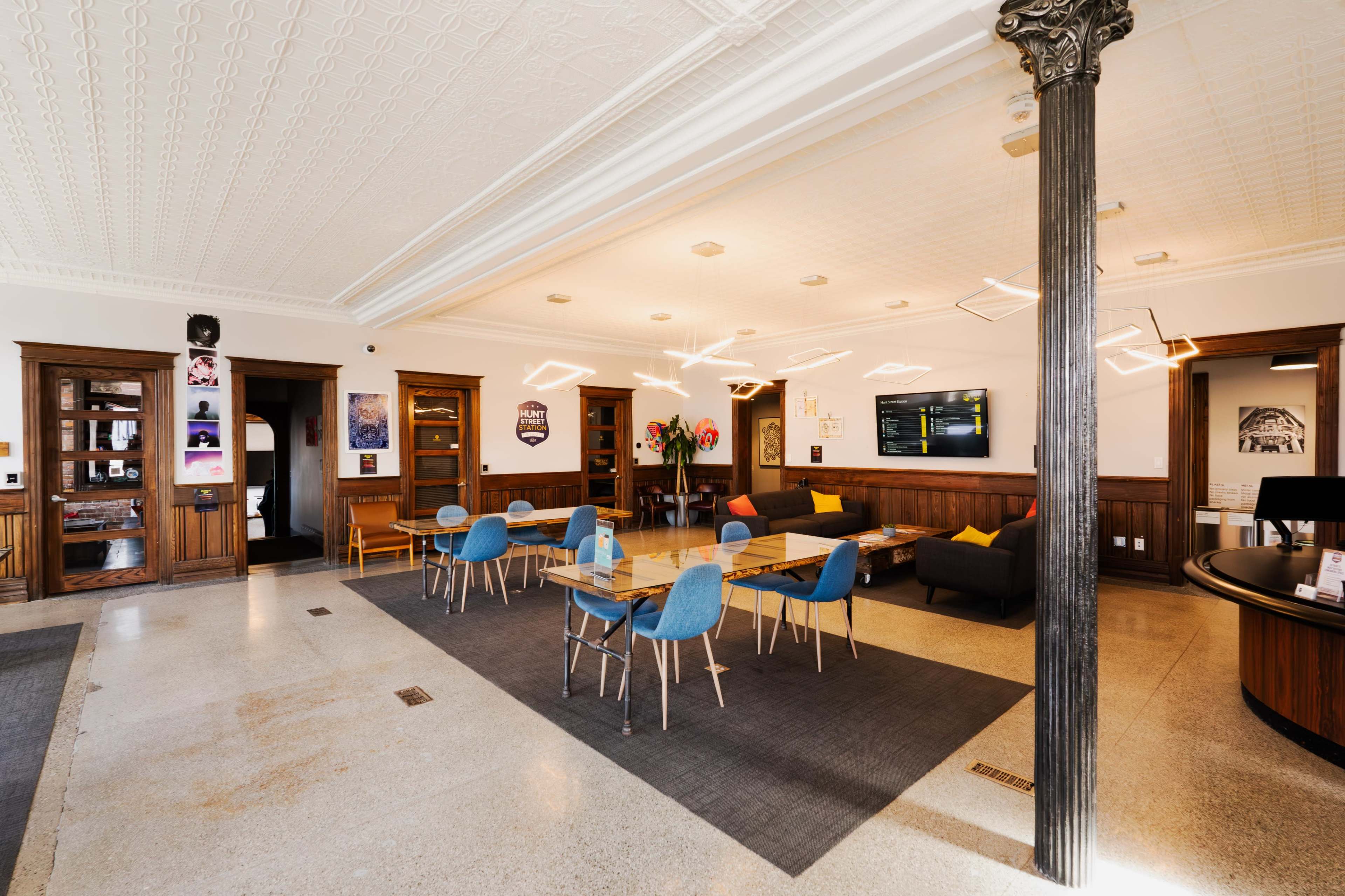 A modern lobby with a seating area, a communal table with blue chairs, and a reception desk, featuring decorative lighting and wooden accents.
