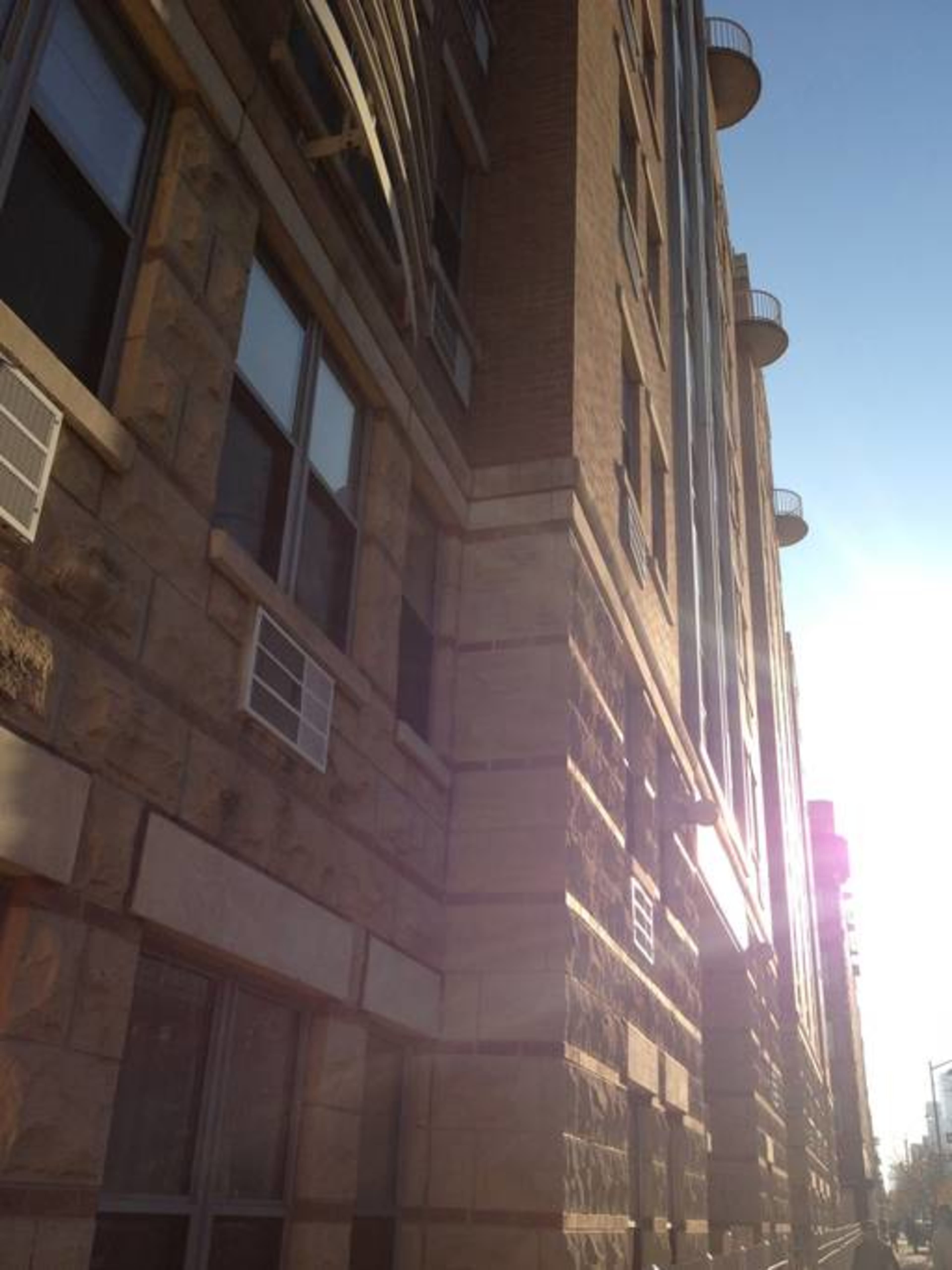 The side of a multi-story building with stone and brick exterior, featuring several windows and air conditioning units.